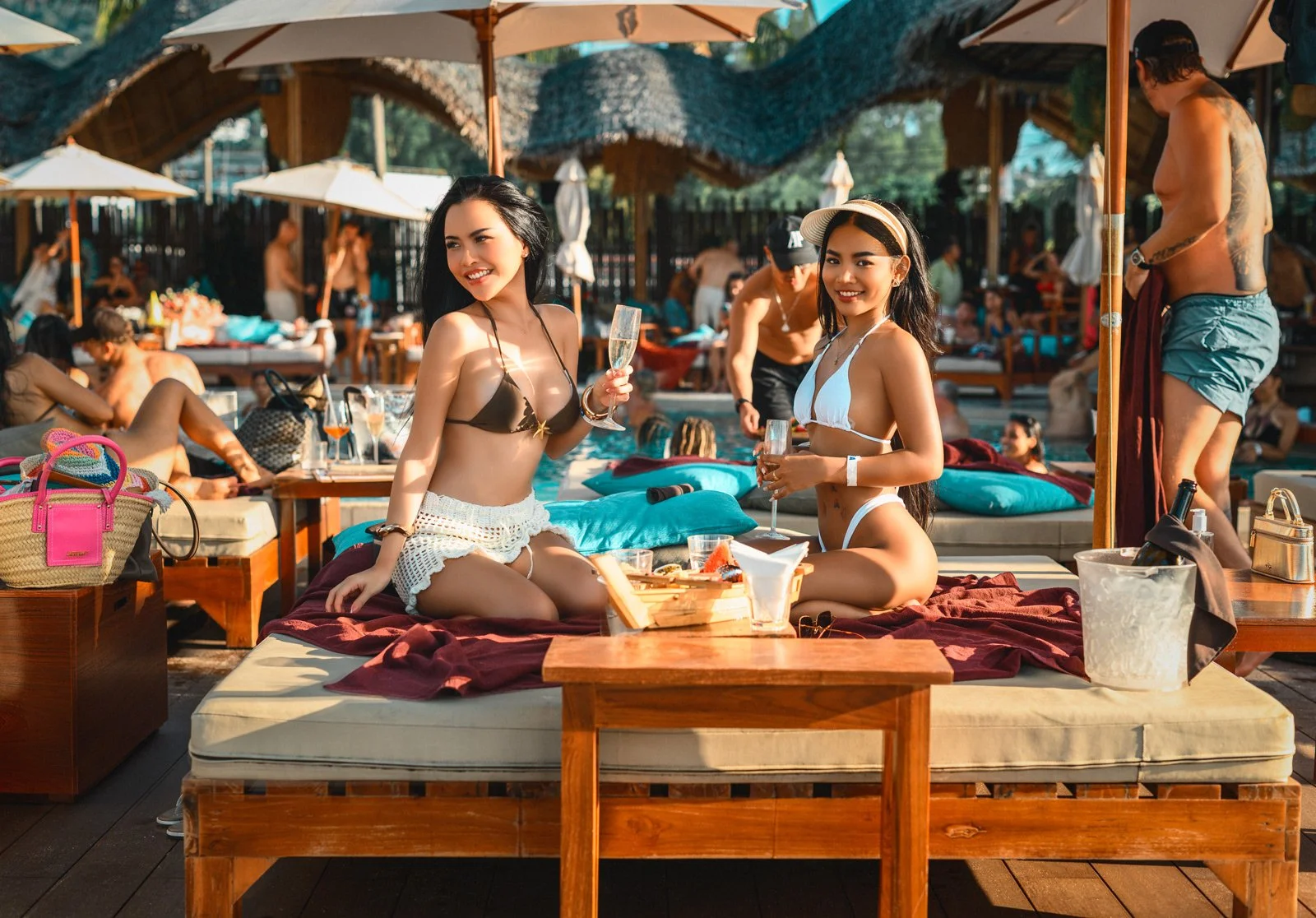 Two women in bikinis sitting on a poolside lounge bed with drinks, surrounded by people relaxing and socializing at a pool party.