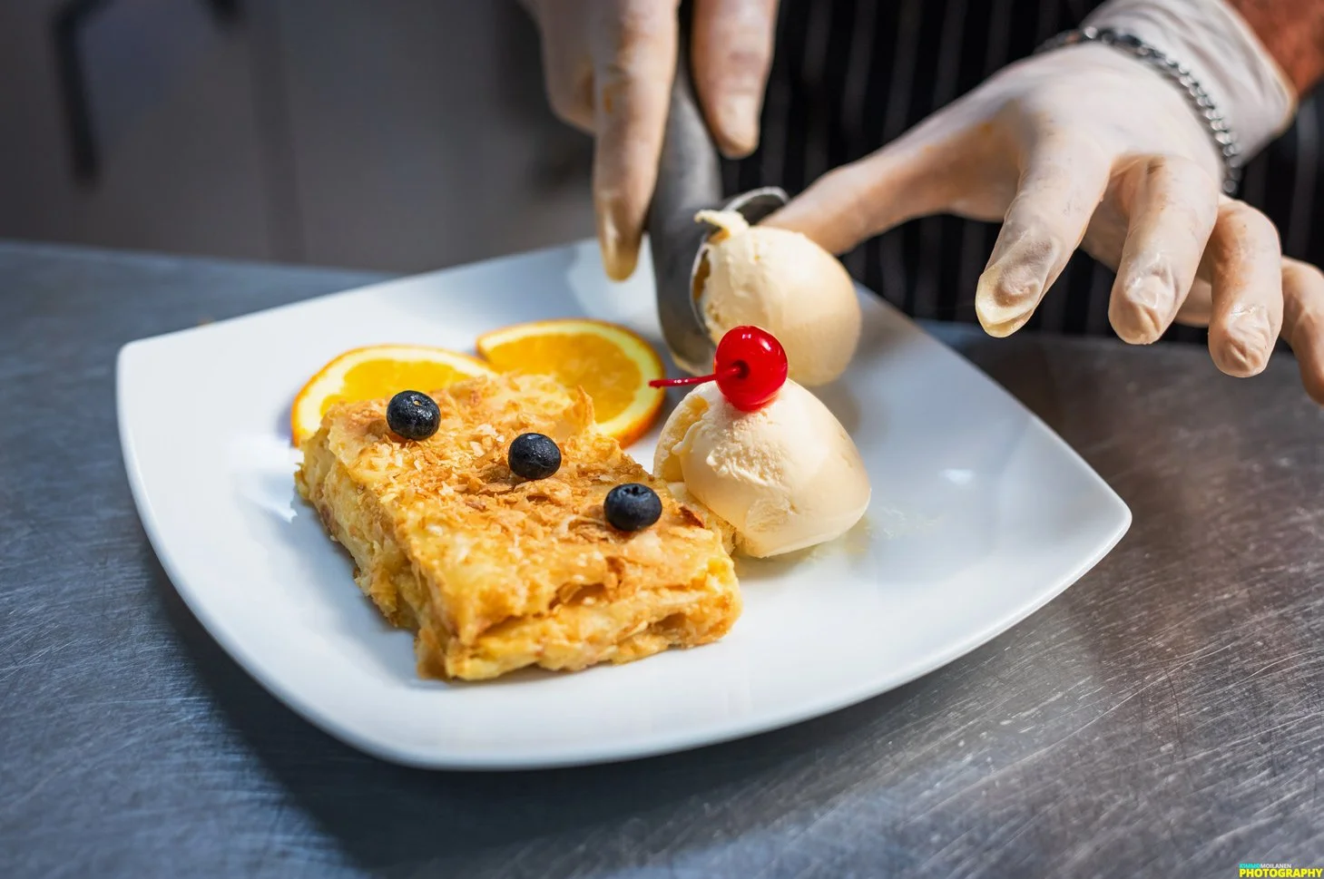 A plate of dessert with slices of orange, scoops of vanilla ice cream topped with a cherry, and blueberry garnishes, being prepared by a person wearing gloves.