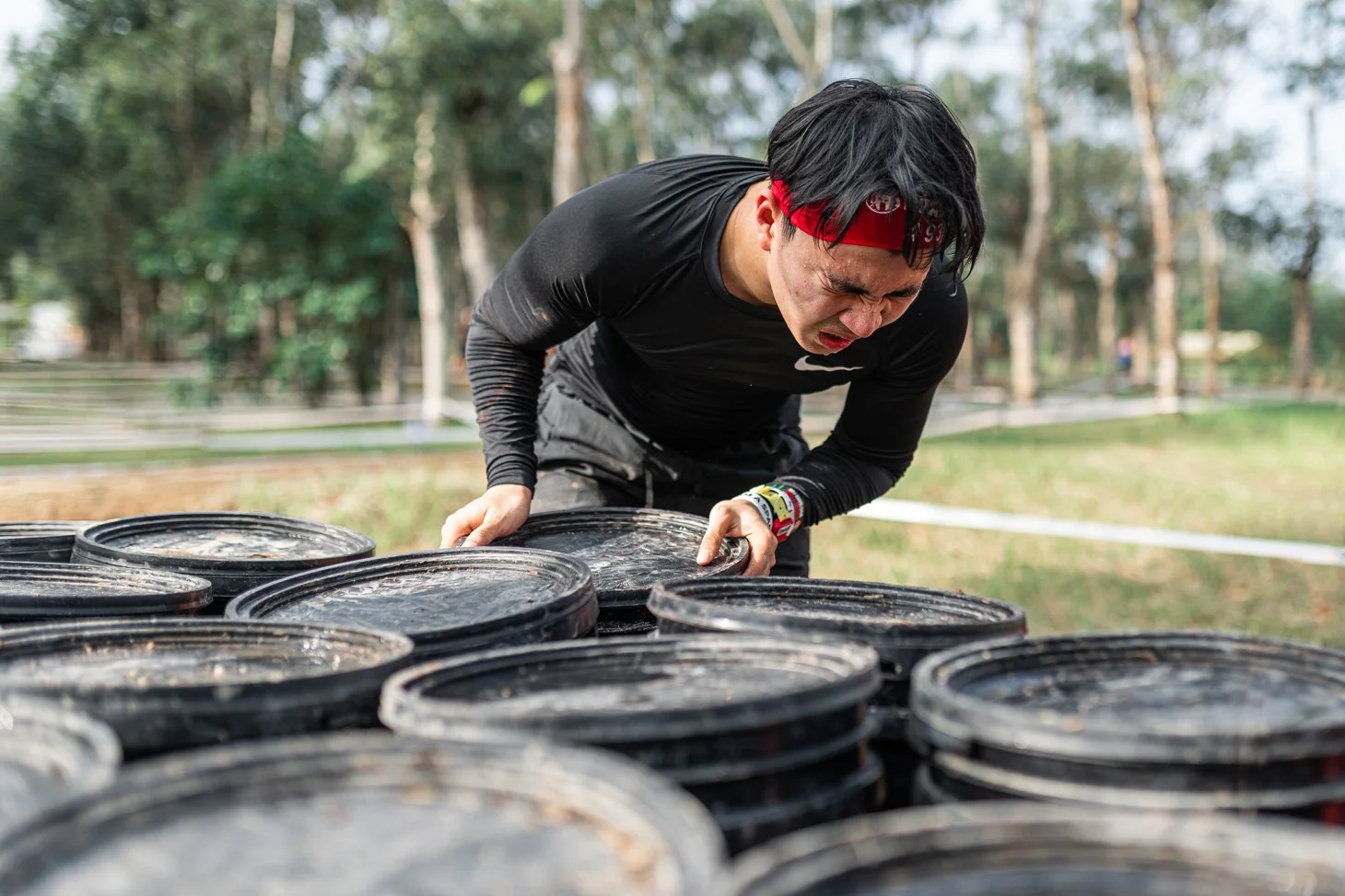 Man with red headband lifting a black plastic bucket outdoors with trees in the background.