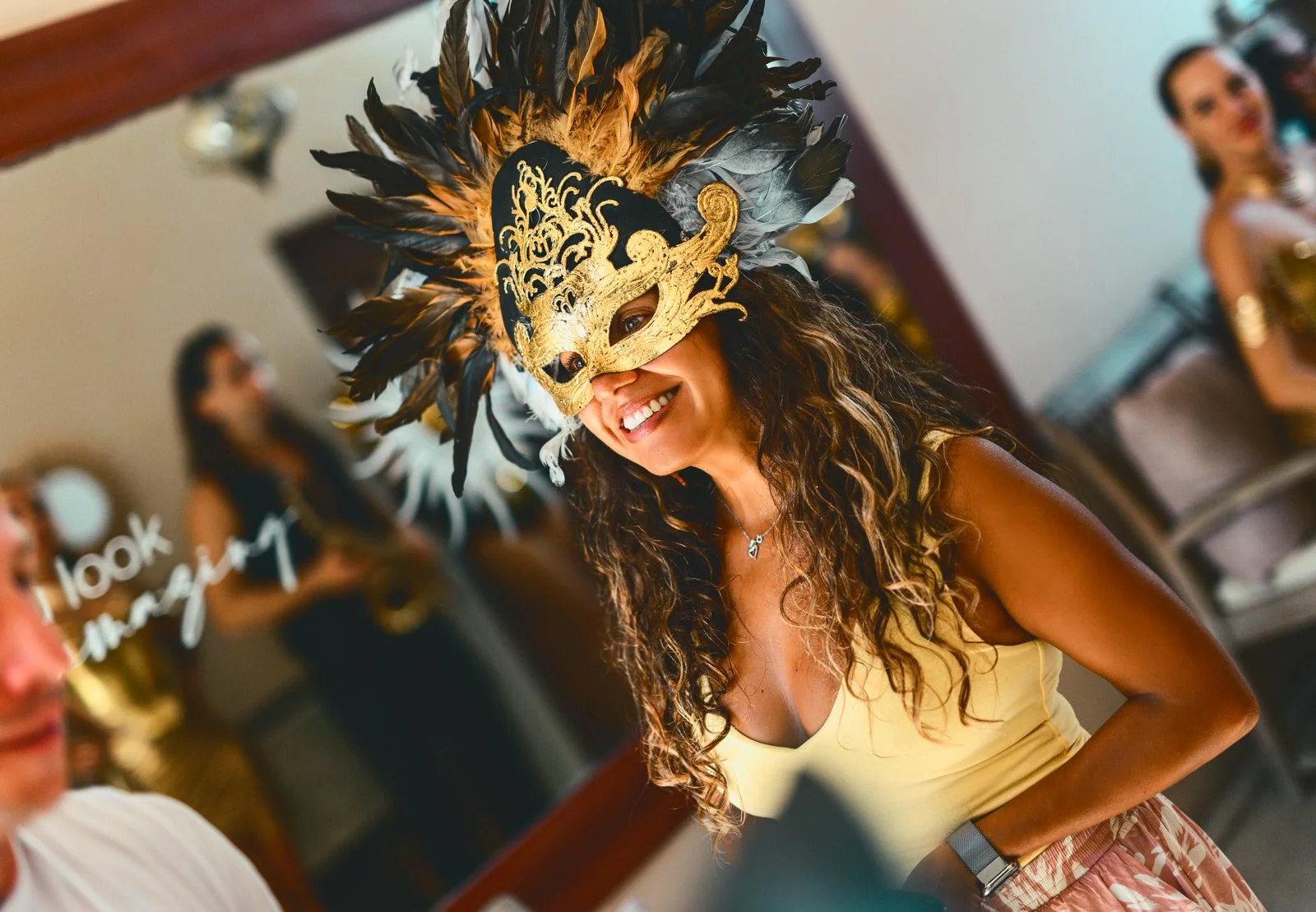 Woman smiling in a costume with a decorative gold and black feathered mask, wearing a yellow top at a festive event.