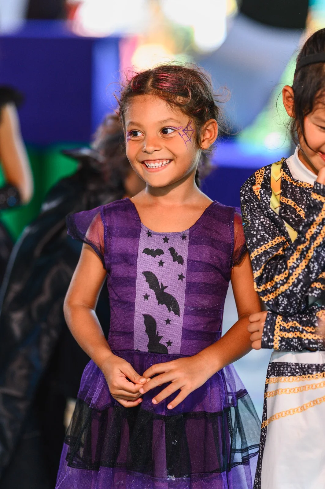 A young girl with a face painting of a purple spider web, smiling, wearing a purple dress with bat and star designs, standing among others dressed in costumes at what appears to be a Halloween celebration.