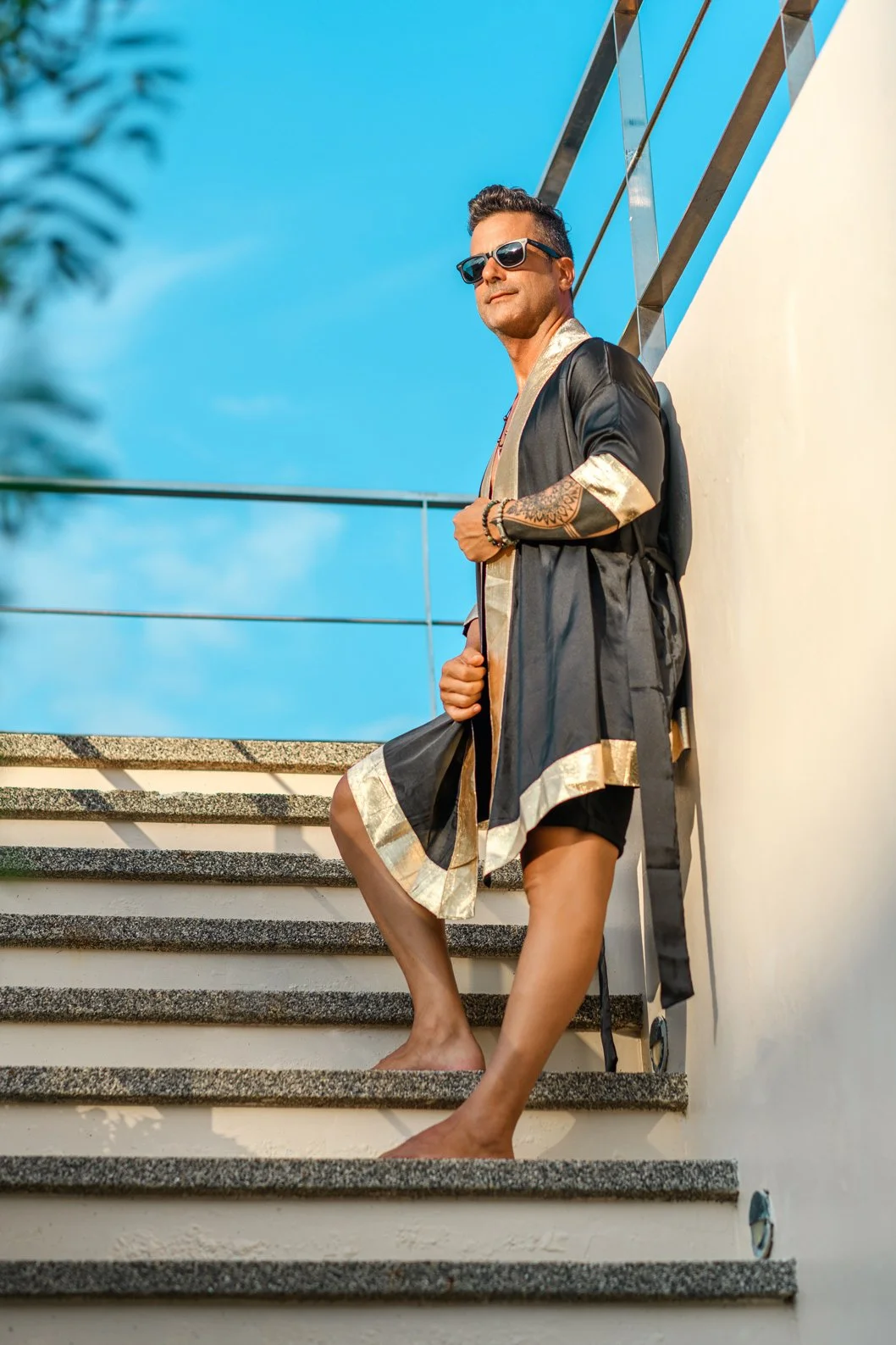 A man standing on outdoor stairs wearing sunglasses, a black and gold robe, and black shorts, with a tattoo on his arm, against a blue sky with a white wall and metal railing.