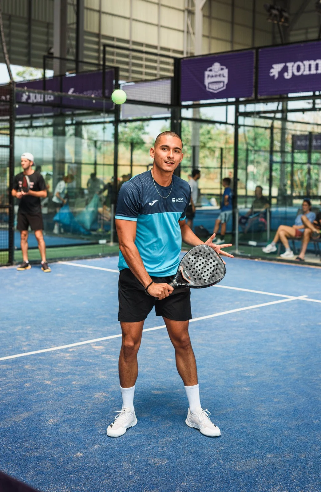 Young man in sportswear holding a racket on an indoor padel court, with other players and spectators in the background.