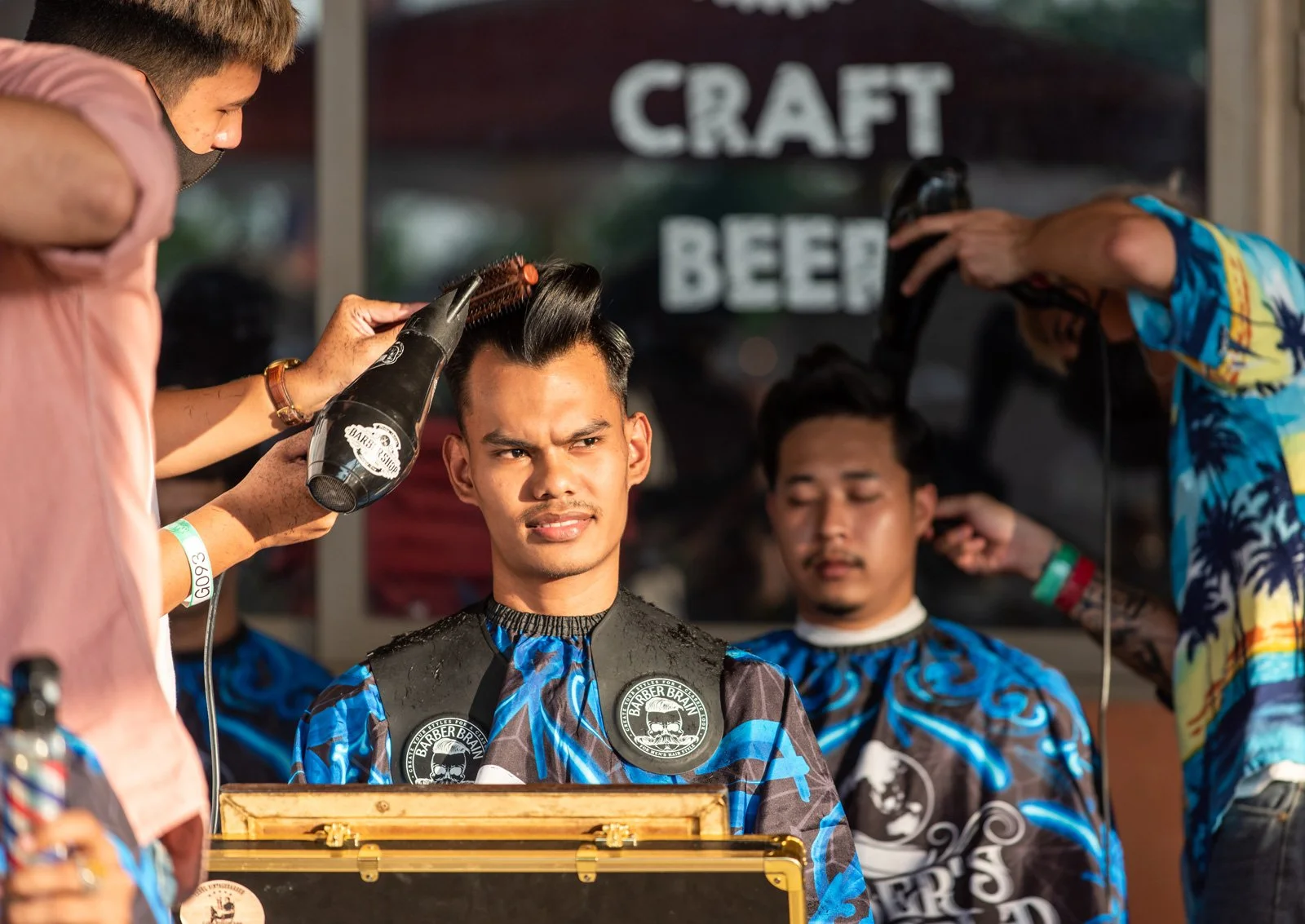 Barber shop with young man receiving a haircut and style, barber using hairdryer, and customer in background holding a hairdryer, with a sign reading 'Craft Beer' in the window.