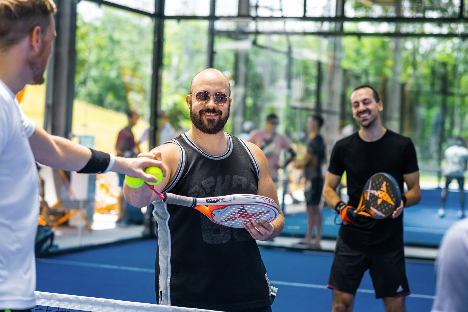 Two men playing pickleball indoors with a glass wall and people in the background. One man is holding a pickleball paddle and is about to hit a green pickleball. The man in the middle is wearing sunglasses and a sleeveless sports shirt, smiling.