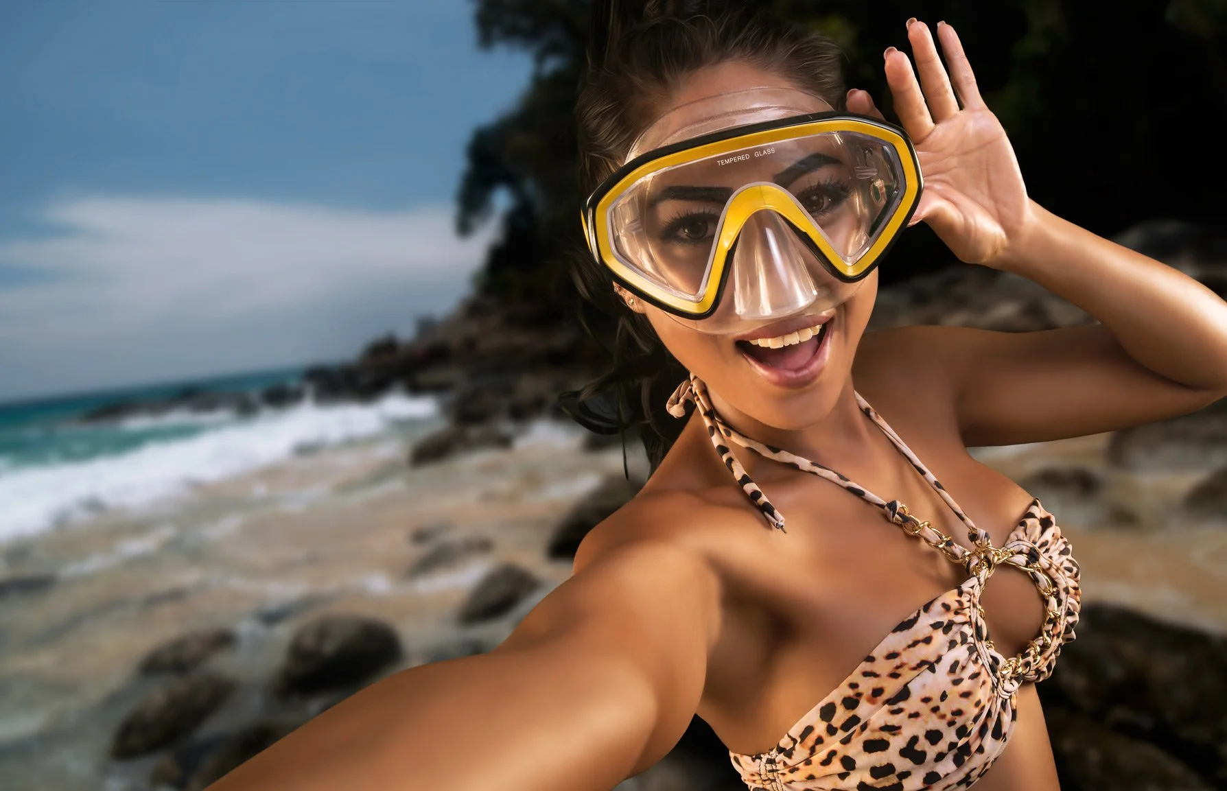 A woman in a leopard print swimsuit taking a selfie on a rocky beach, wearing a yellow snorkeling mask, with blue sky, ocean waves, and rocks in the background.
