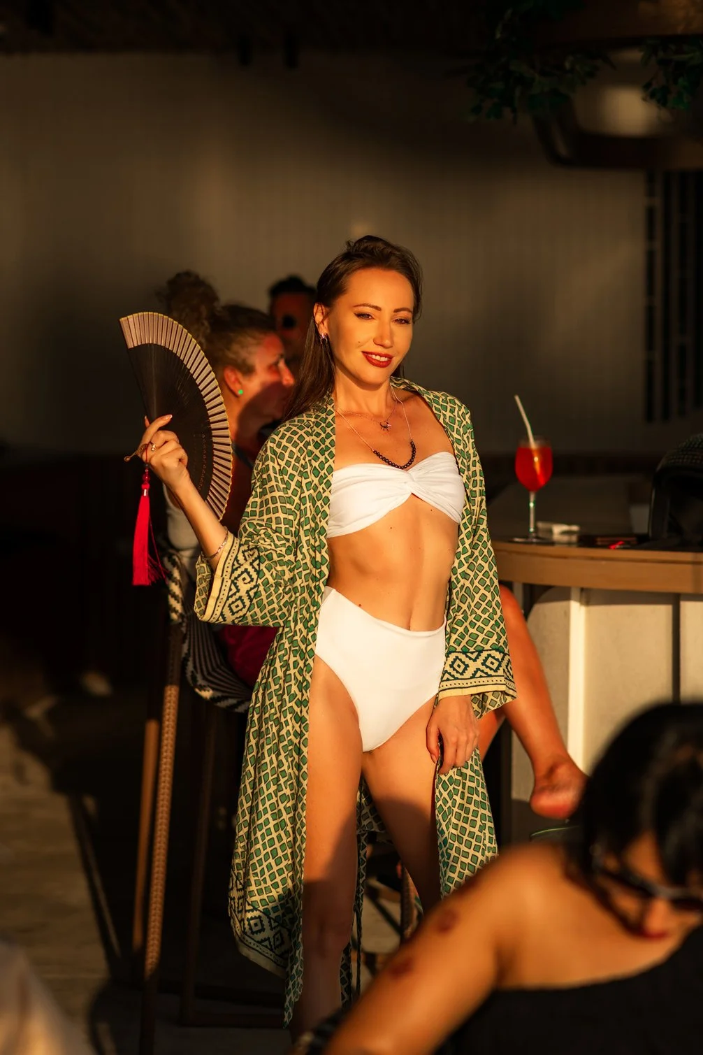 A woman in a white bikini and patterned kimono holding a fan at a social gathering or party.
