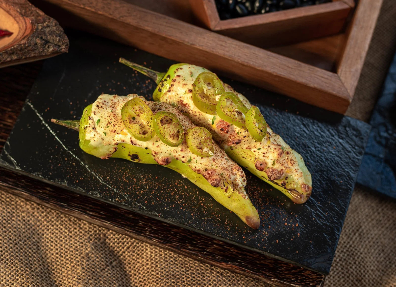 Two stuffed green peppers topped with sliced green chilies, spices, and herbs on a black slate serving board.
