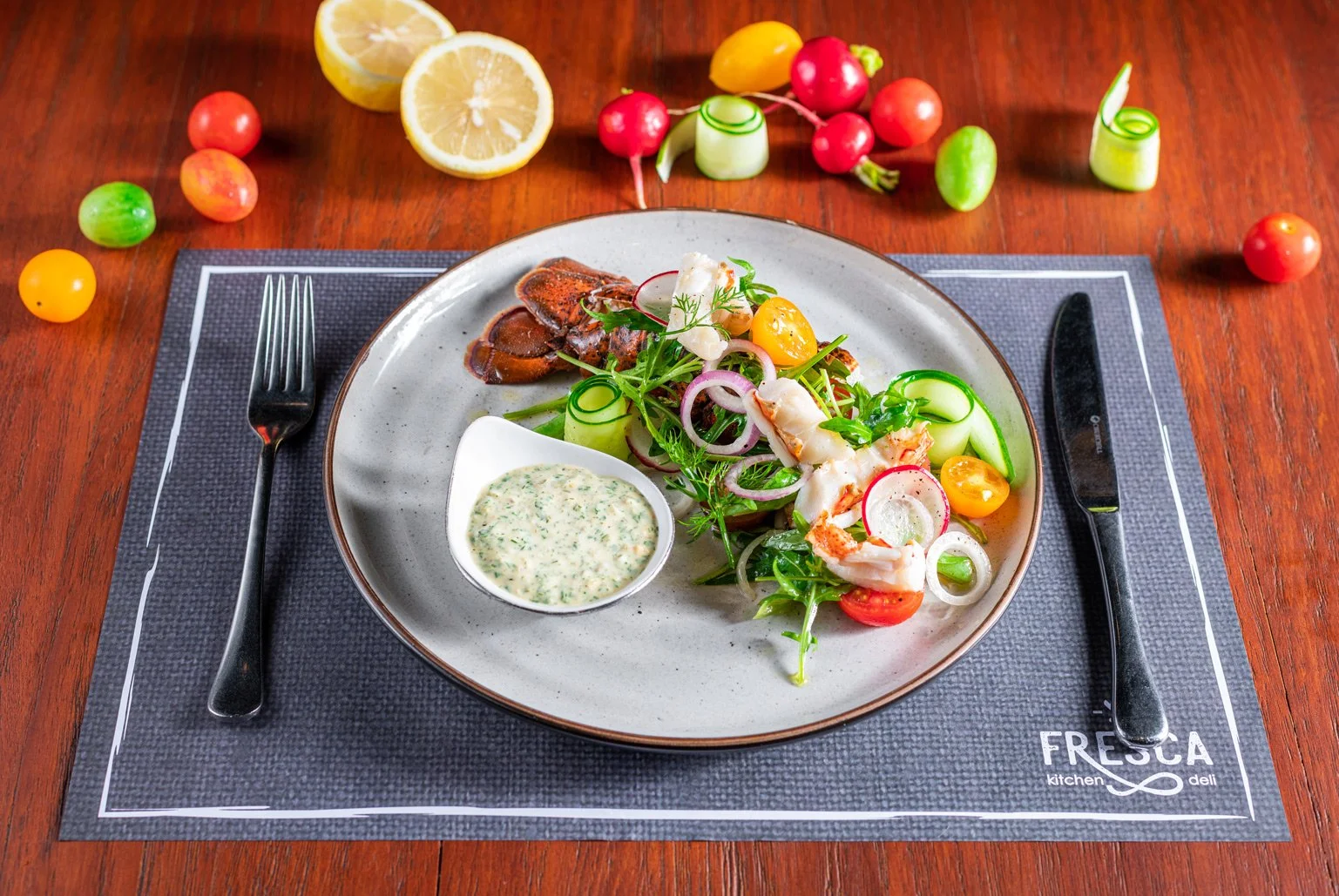 A plate of seafood salad with vegetables, sliced radishes, cucumber, cherry tomatoes, greens, and a side of creamy herb sauce on a gray placemat on a wooden table, surrounded by lemon halves and colorful cherry tomatoes.