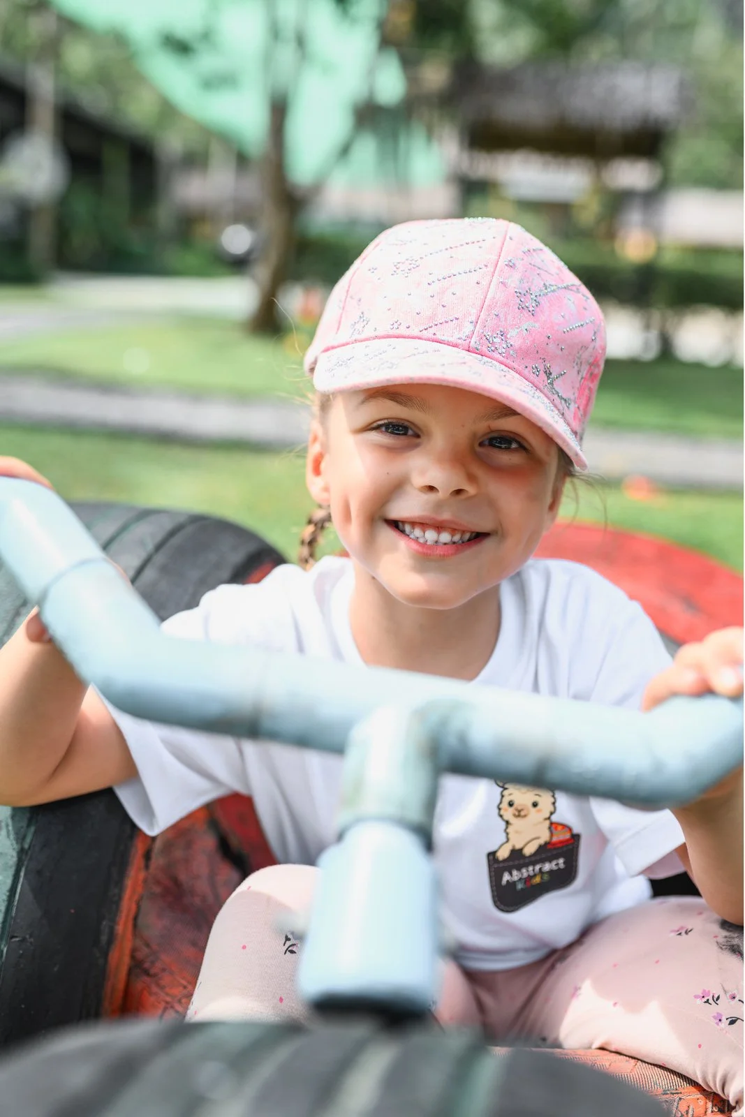 A young girl with a pink cap and white shirt smiling while sitting on a playground tire swing.