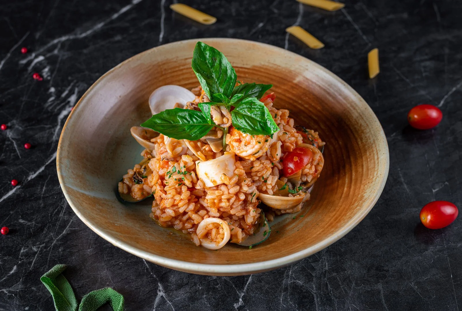 A bowl of seafood risotto garnished with fresh basil leaves, served on a black marble surface with scattered cherry tomatoes and penne pasta.