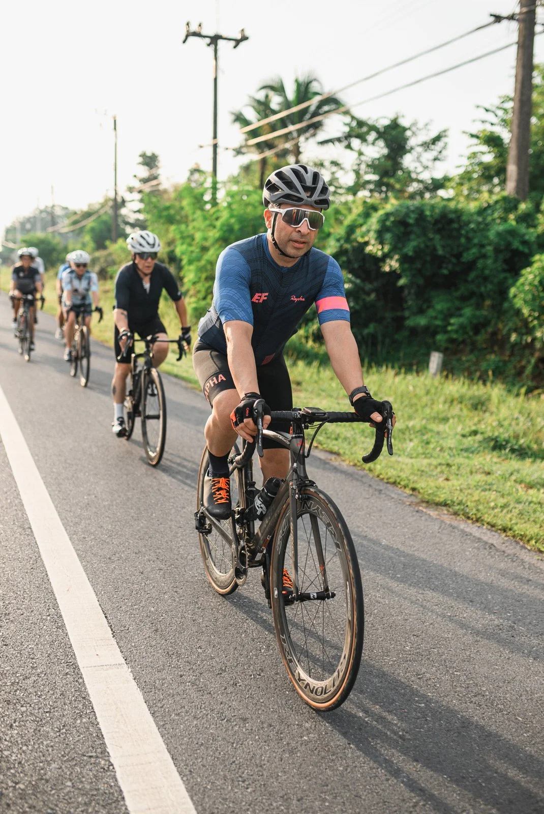 A group of cyclists riding on a paved road surrounded by greenery and power lines, with the lead cyclist wearing sunglasses and a blue cycling jersey.