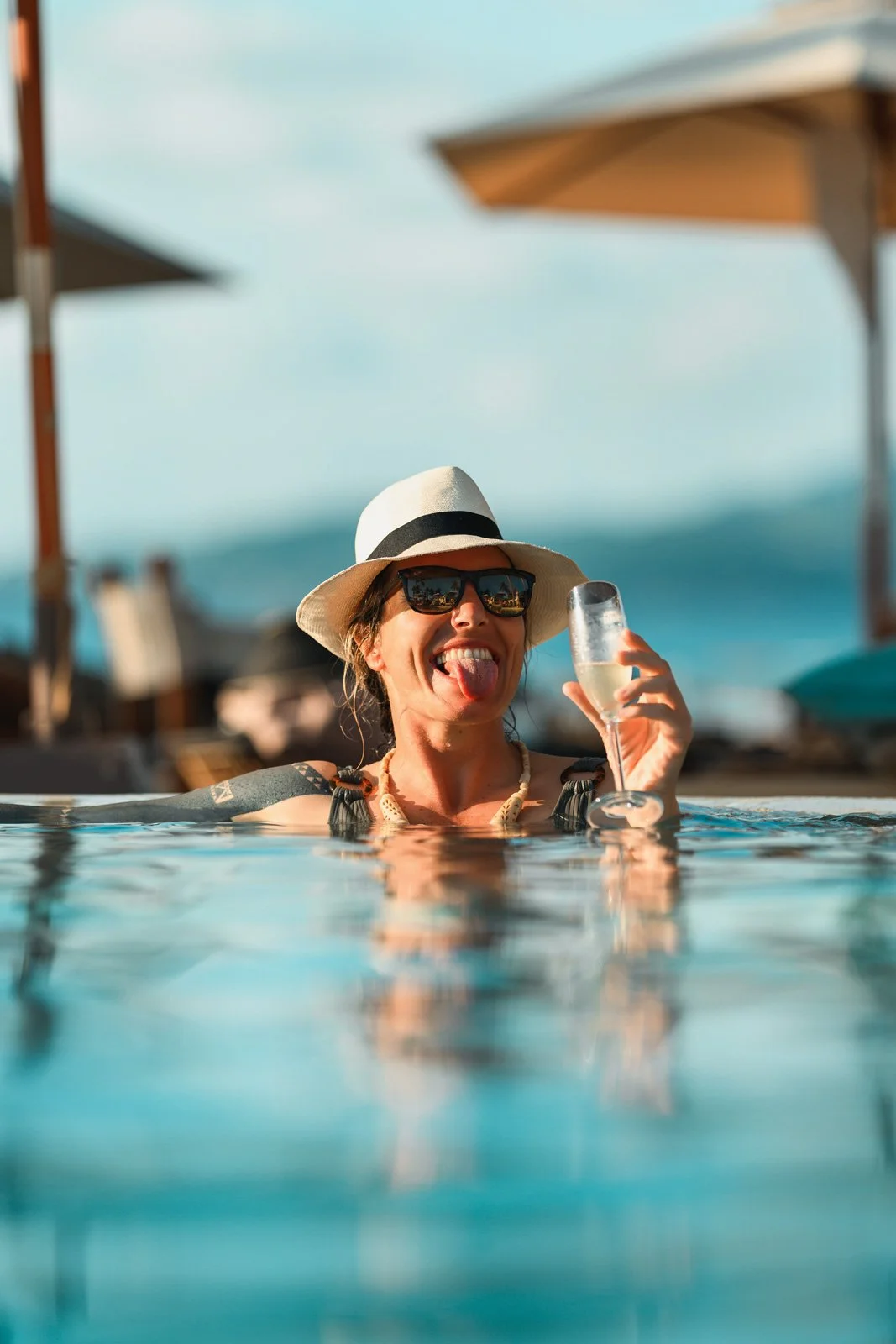 A woman in a swimming pool wearing sunglasses, a wide-brimmed hat, and a necklace, holding a glass of champagne and sticking out her tongue.