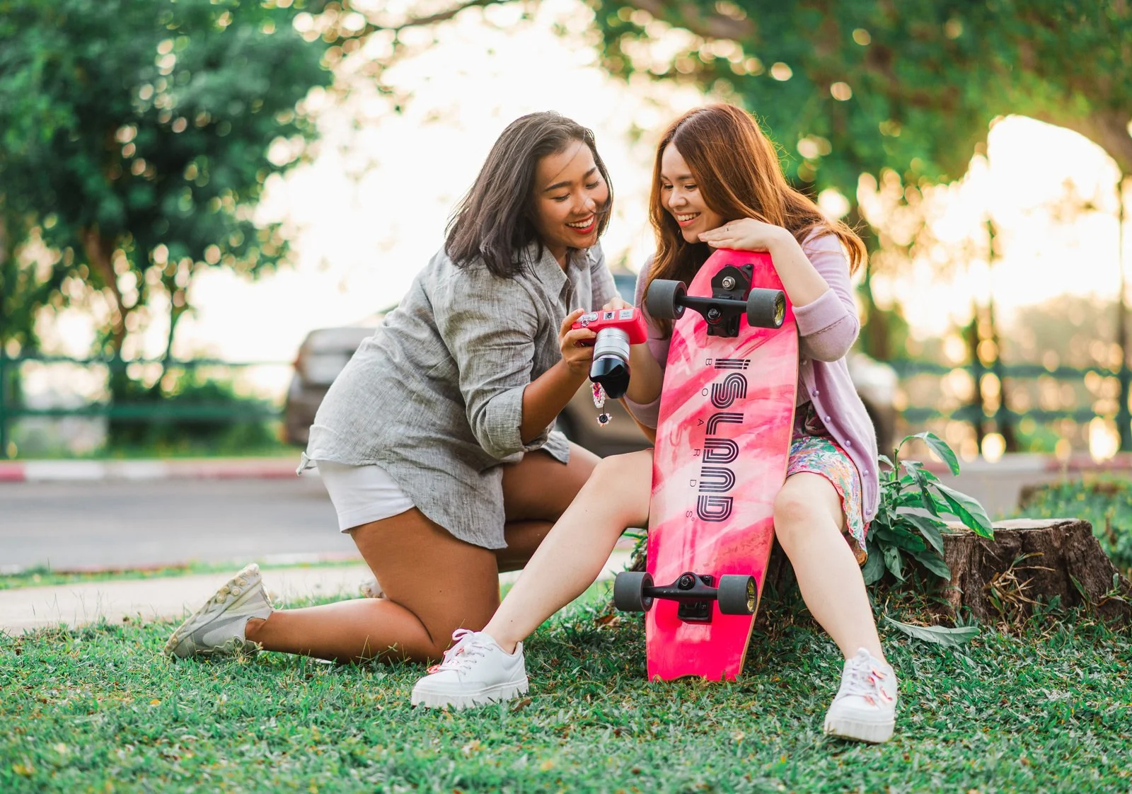 Two young women outdoors, one showing a camera to the other with a skateboard nearby, sitting on grass near a tree.