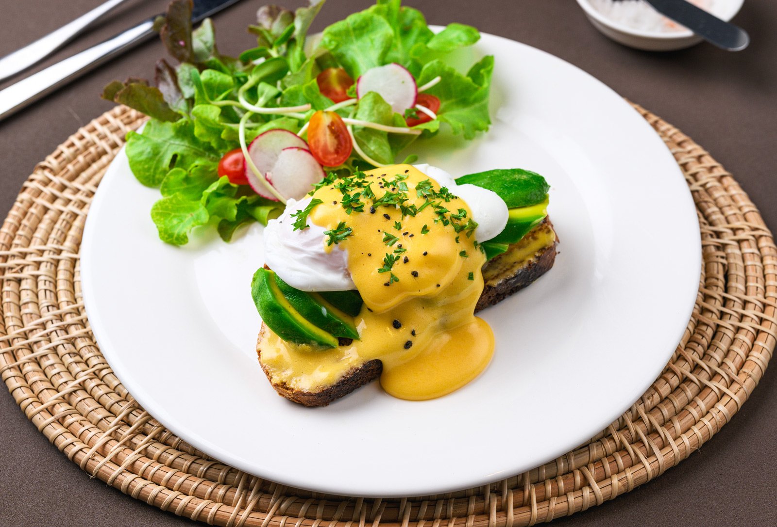 A plate with eggs benedict topped with hollandaise sauce, served with a fresh vegetable salad on the side, all on a woven placemat.