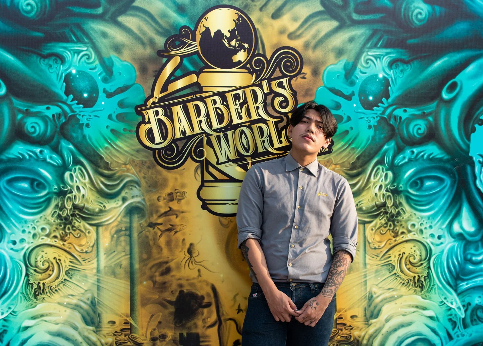 A young man standing in front of a vibrant, colorful mural that has a large illustration of a light bulb with the words "Barbers World" and a globe on top. The mural features swirling blue and yellow abstract patterns.