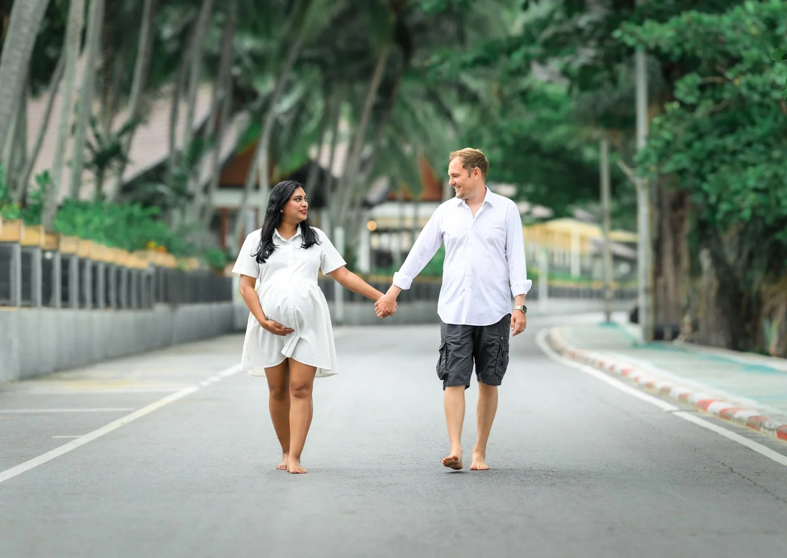 Pregnant woman and man walking together barefoot on a street, holding hands, with trees and houses in the background.