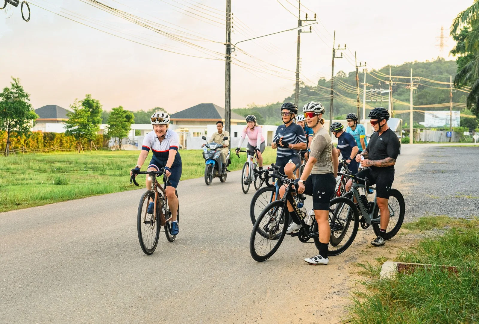 Group of people cycling outdoors on a scenic road during late afternoon, with some taking a break and chatting, in a suburban area with houses, trees, and power lines visible.