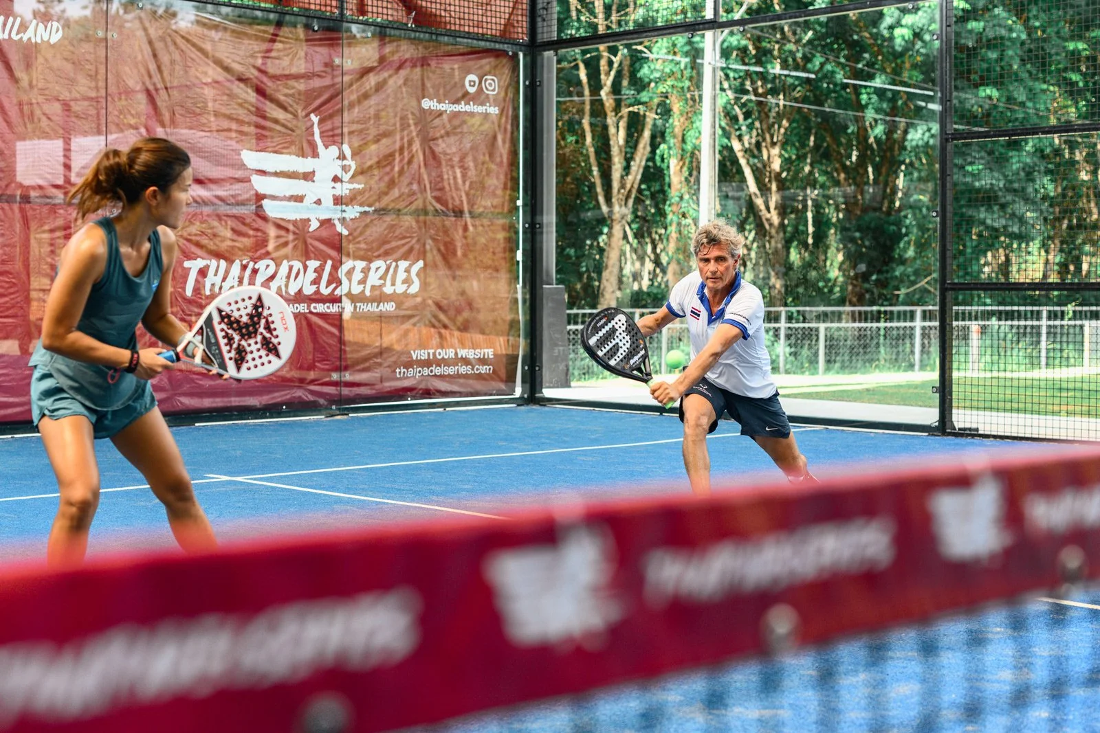 Two people playing padel tennis on an outdoor court with a red banner that says "Thailand Series" in the background. The man is hitting the ball with a paddle, while the woman is preparing to return it.