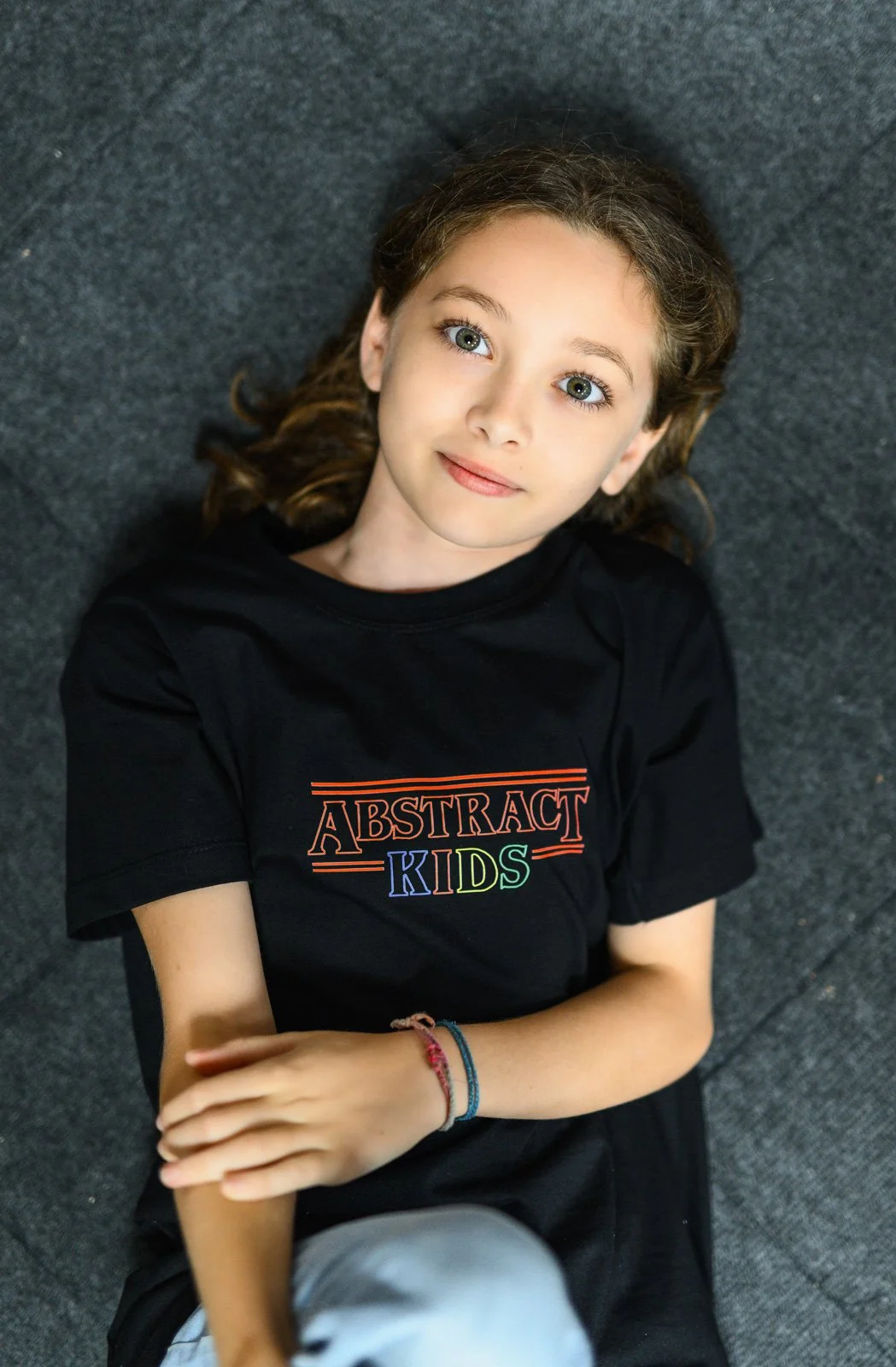 A young girl with curly hair and blue eyes is lying on a dark, textured floor, wearing a black T-shirt that says "ABSTRACT KIDS" in colorful letters.
