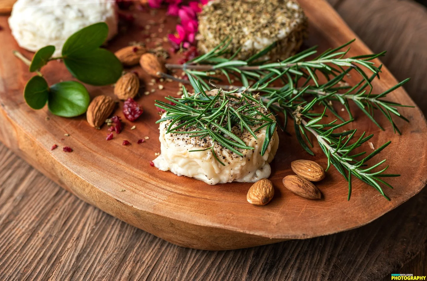 Herb and cheese platter on wooden serving board featuring rosemary sprigs, almonds, green leaves, and cheese with herbs.