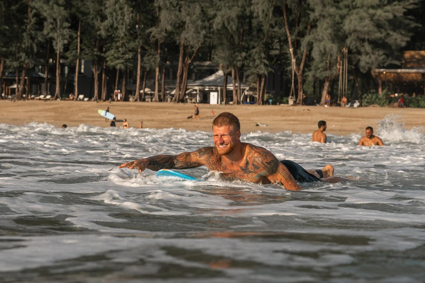 A man with tattoos riding a surfboard in the ocean, smiling, with a beach and trees in the background.