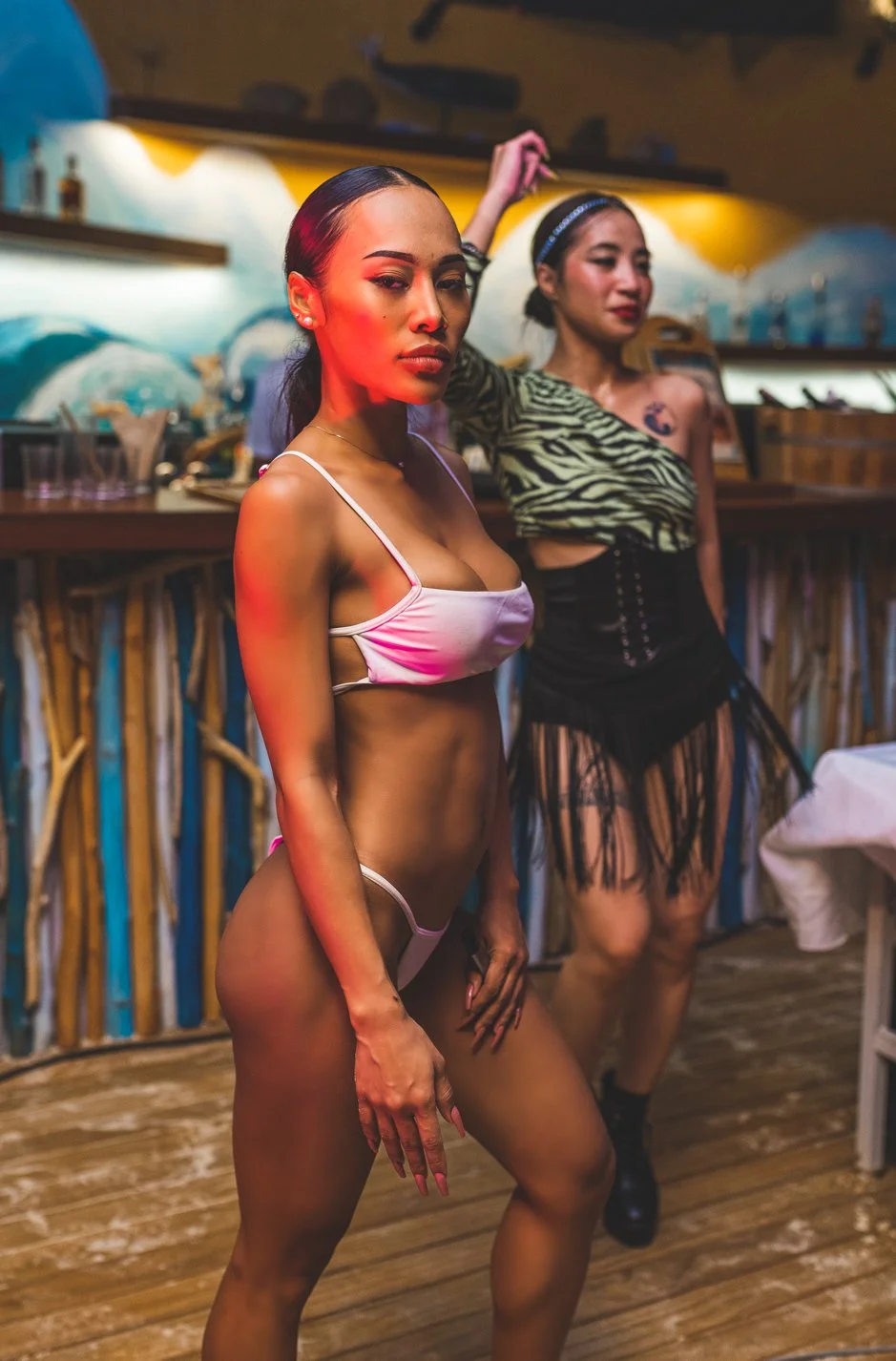 Two women in revealing outfits posing indoors; one in a satin pink and white top and matching underwear, the other in a zebra print top and black fringe skirt.