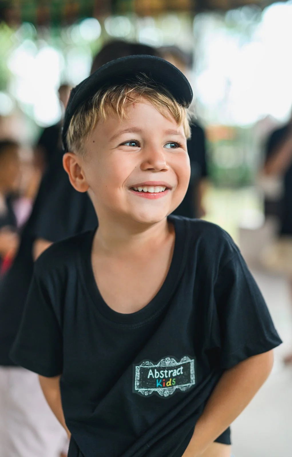 Smiling young boy with blond hair wearing a black cap and black t-shirt labeled "Abstract Kids," indoors with other children and blurred background.