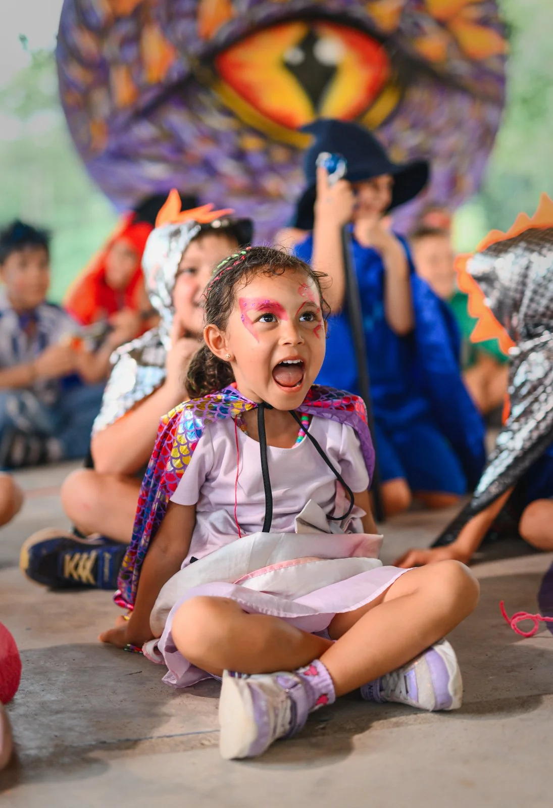 Children participating in a festive event wearing colorful costumes, with one girl in the foreground wearing face paint and a cape, sitting on the ground with her mouth open, and a large lion face mask or decoration in the background.