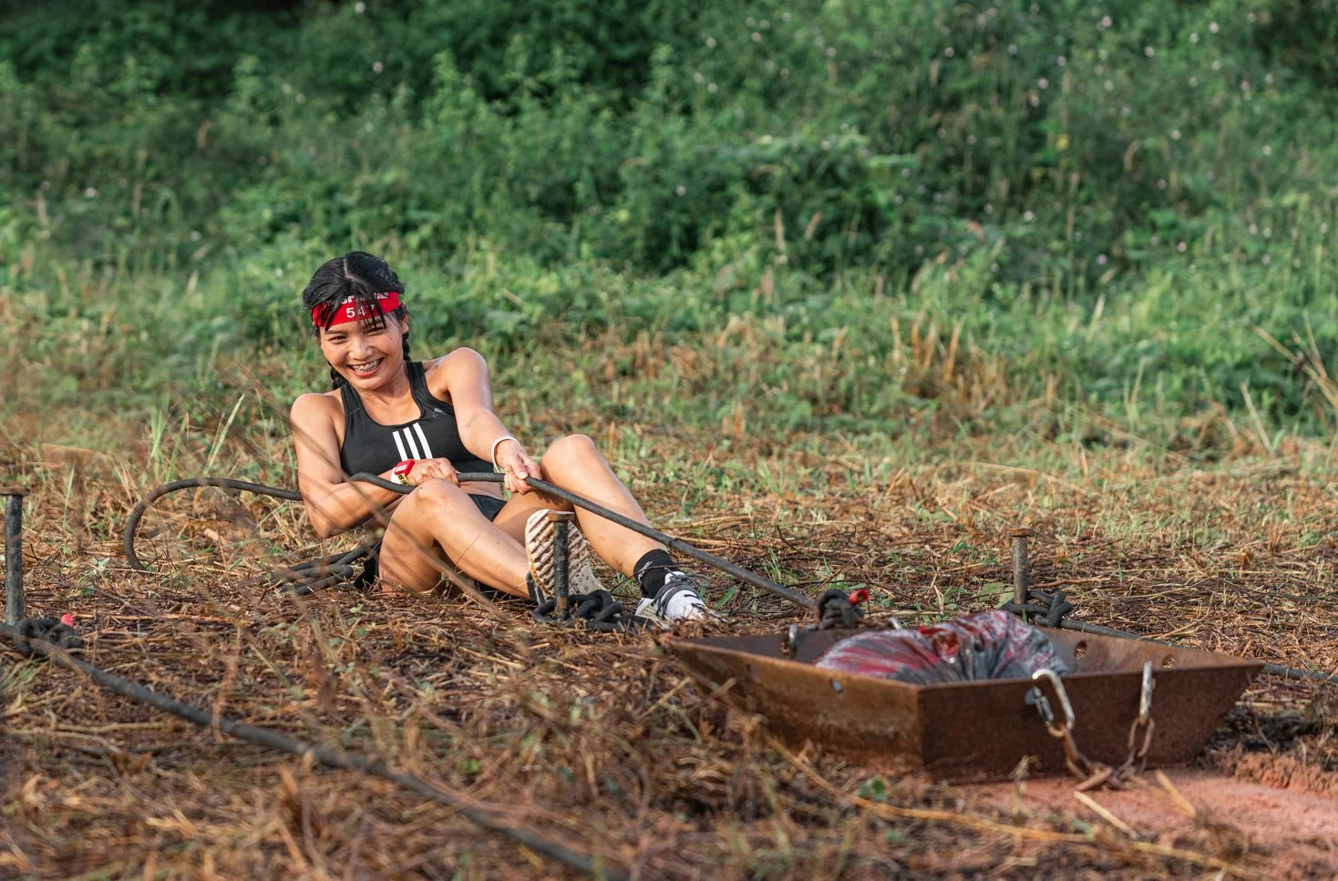 A young woman in athletic clothing is sitting on the ground and smiling, holding onto some ropes attached to a sled with a bag on it, in an outdoor area with grass and dirt.