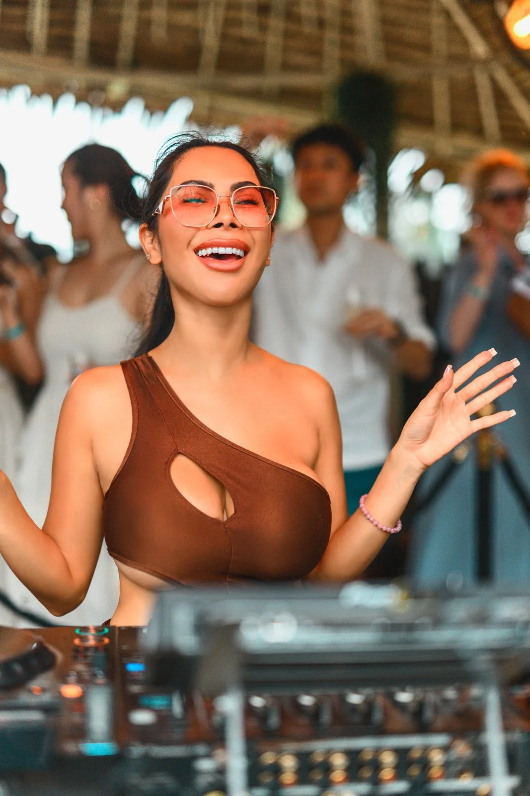A woman with sunglasses smiling and dancing at a party with a DJ setup in front of her, surrounded by other people.
