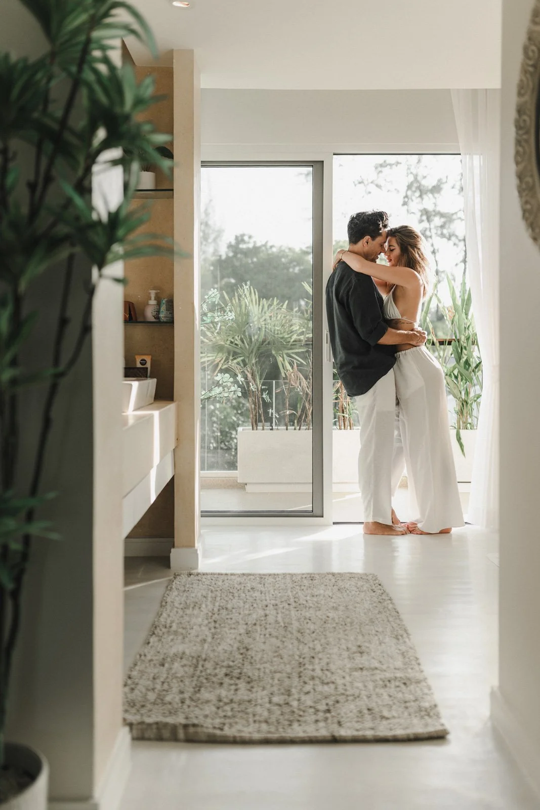 A couple is embracing and kissing in a bright room by a large glass sliding door with potted plants outside.