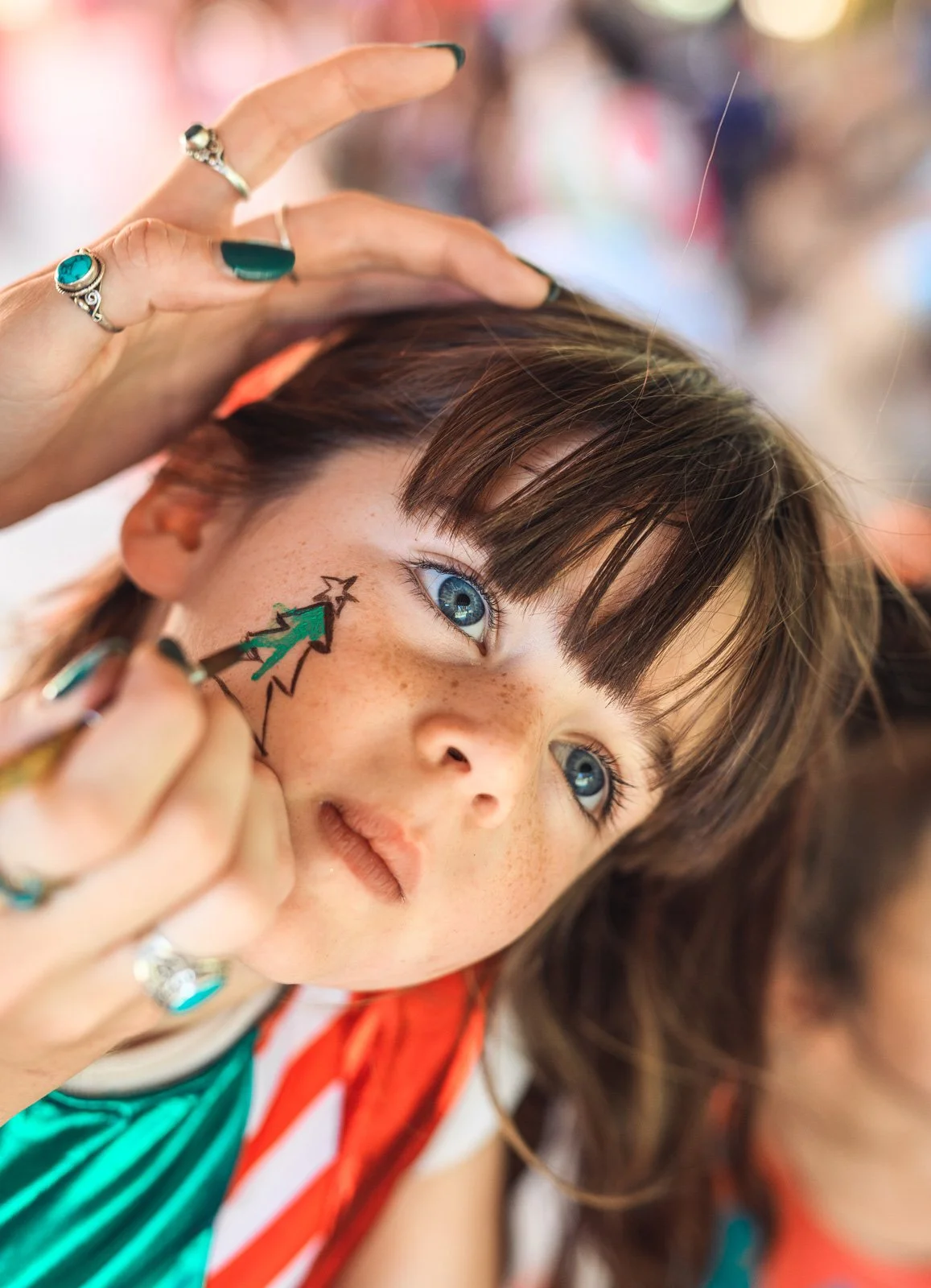 A young girl with blue eyes and freckles gets her face painted with a green Christmas tree with a yellow star on top. A person with rings on their fingers is doing the face painting. The girl has brown hair and is wearing a striped shirt.