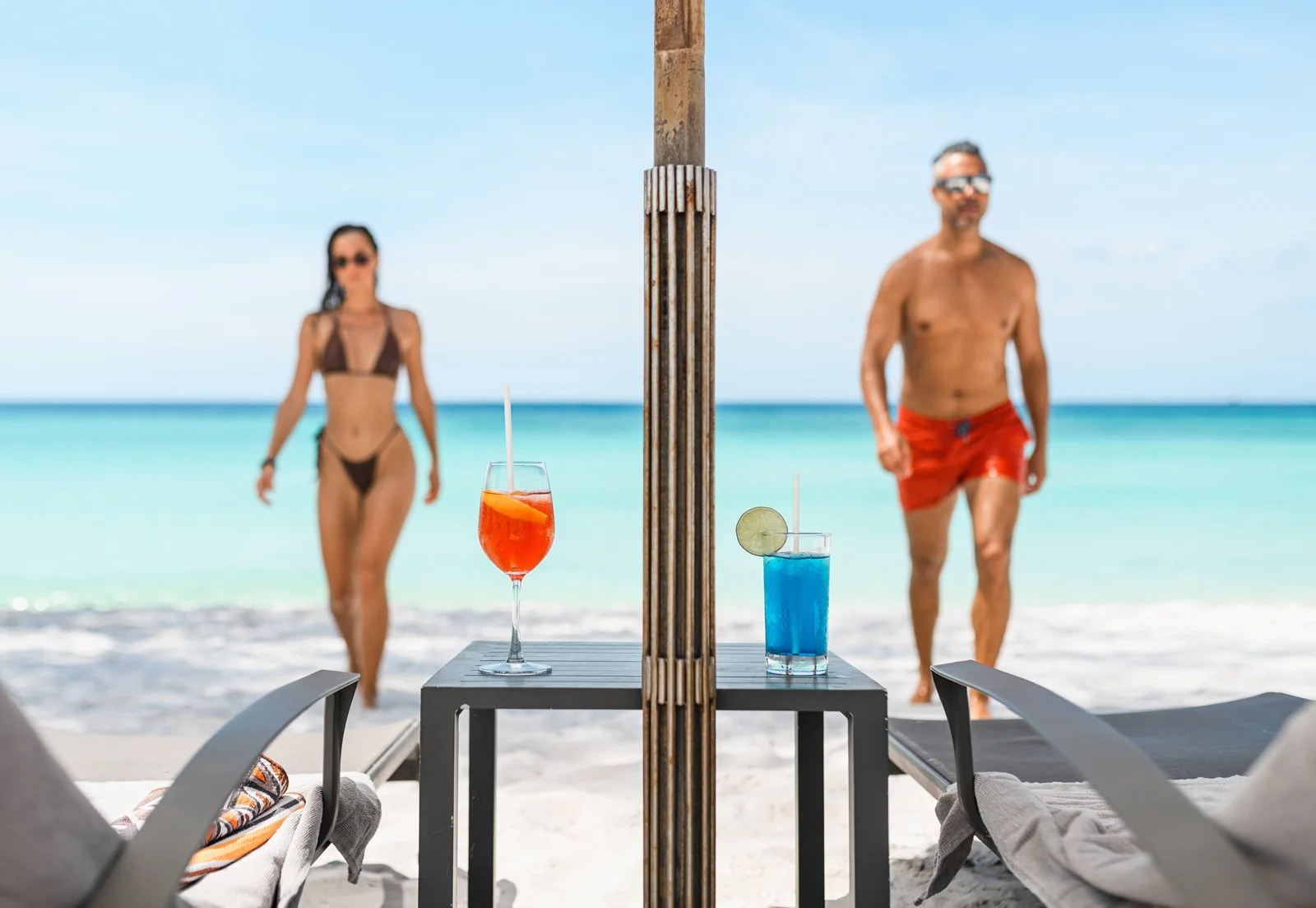 Two colorful cocktails on a table at the beach, with a woman in a bikini and a man in swim trunks walking along the shoreline in the background.