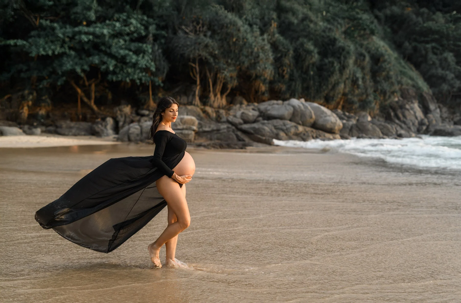 A pregnant woman standing on a beach at sunset, wearing a flowing black dress that drapes on the sand and clouds the shoreline.