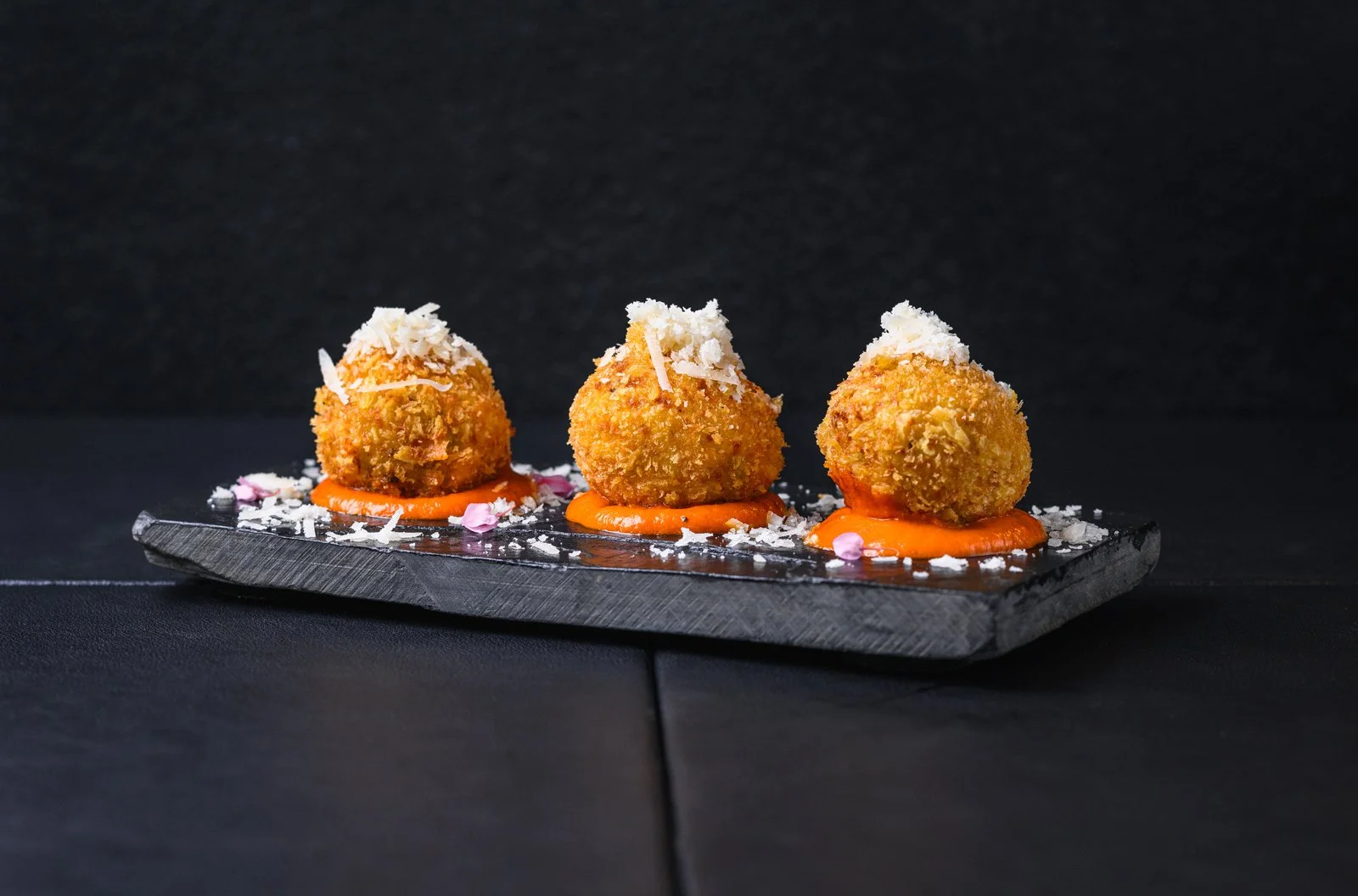 Three fried appetizer balls with bread crumbs on top, served on a black stone slab with orange sauce underneath, garnished with white cheese shavings and pink edible flower petals.