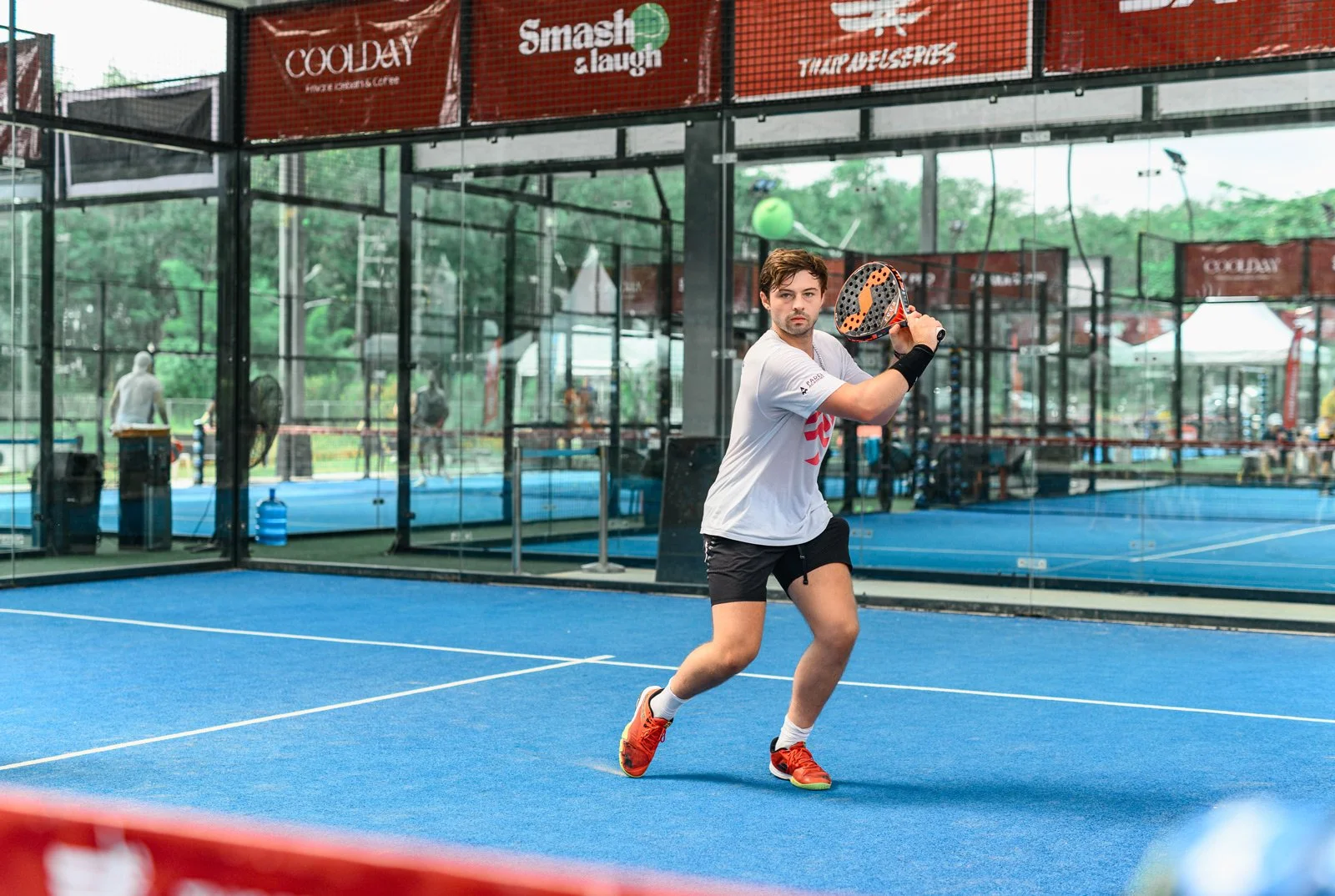A young man playing paddle tennis on an indoor court with blue flooring, wearing a white T-shirt, black shorts, and orange sports shoes, holding a paddle racket in a ready position.