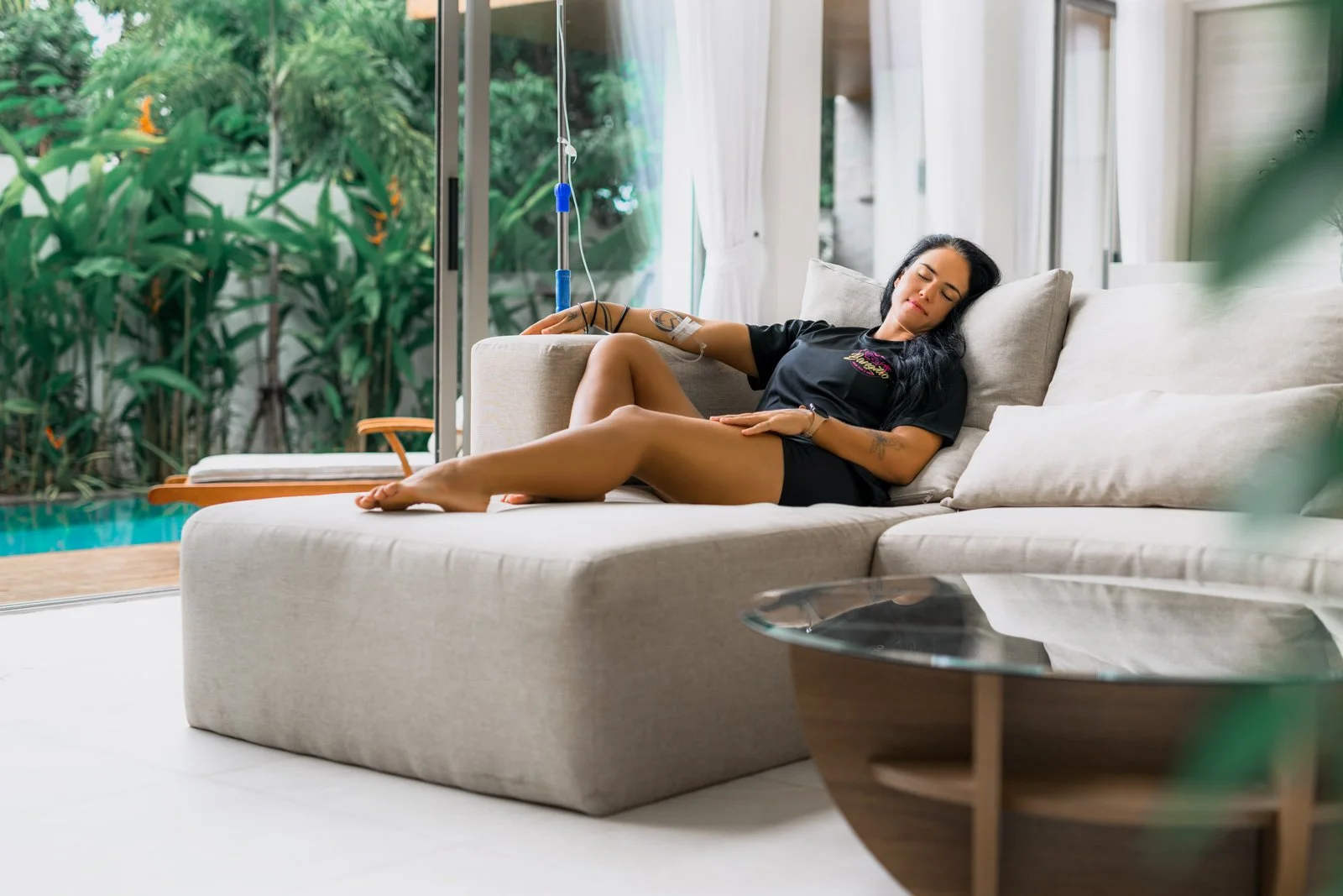 A woman lying on a beige sofa inside a house, with IV drip attached to her arm, her eyes closed, and a peaceful expression, with a view of green foliage outside through large glass doors.