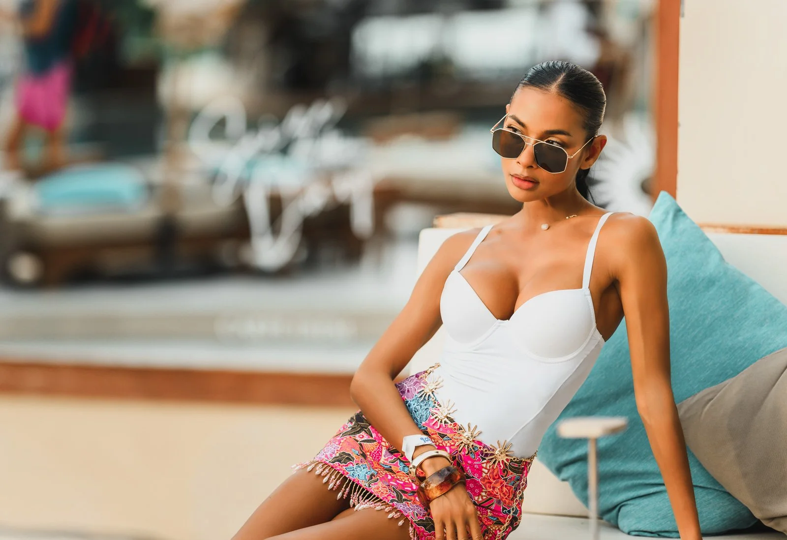 A woman wearing sunglasses, a white top, and colorful patterned shorts sitting on a lounge chair with a turquoise pillow behind her, at what appears to be a resort or poolside location.