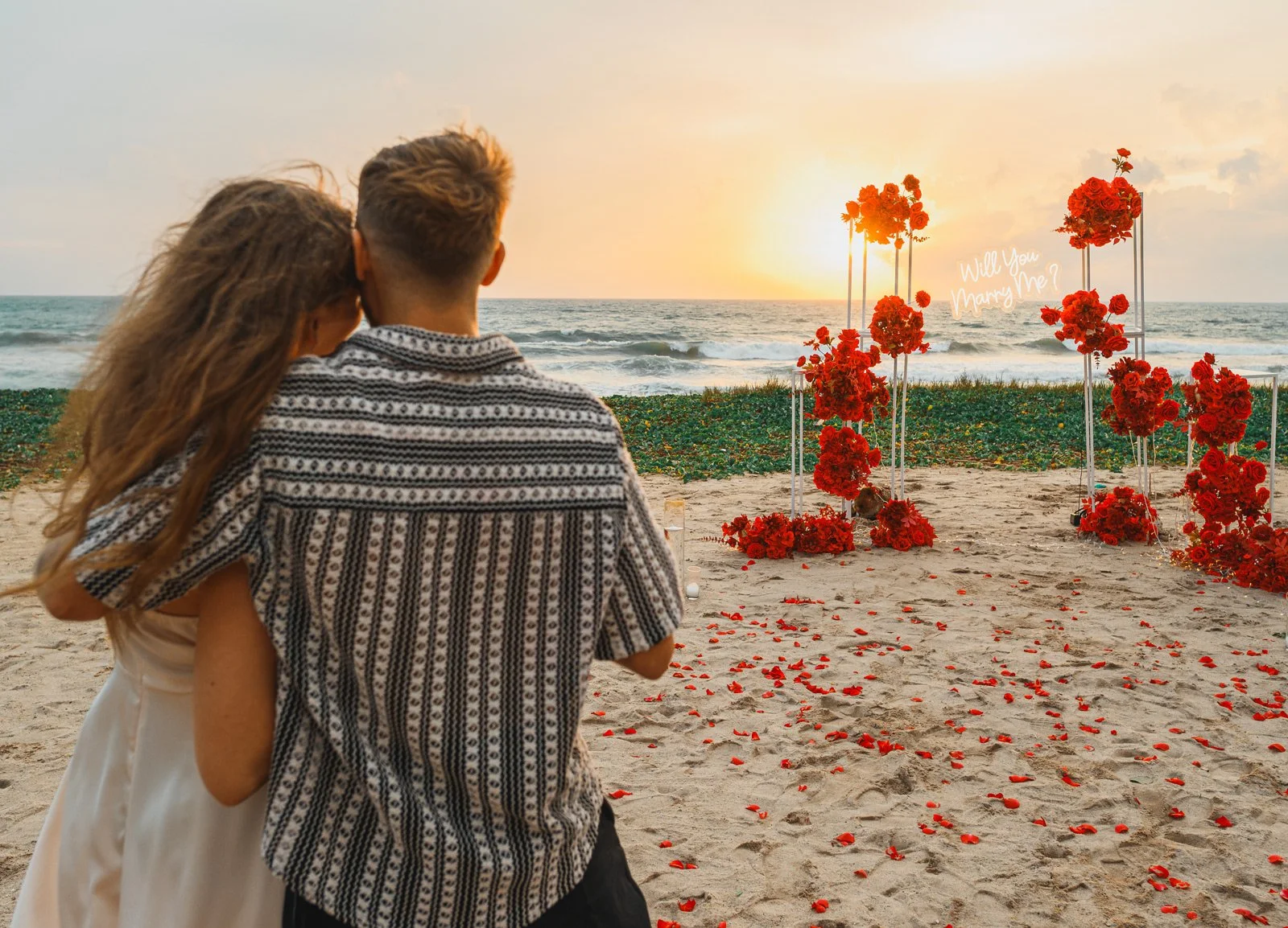 A couple sitting on the beach during sunset, with a floral wedding arch and 'Will You Marry Me?' sign in the background.