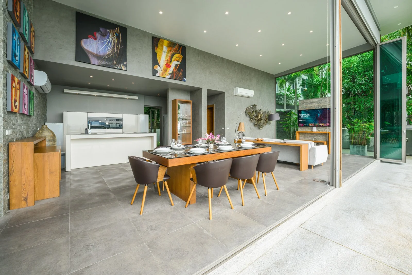 Modern open-concept dining and living area with large glass doors opening to a lush green outdoor space, decorated with contemporary art and furniture.