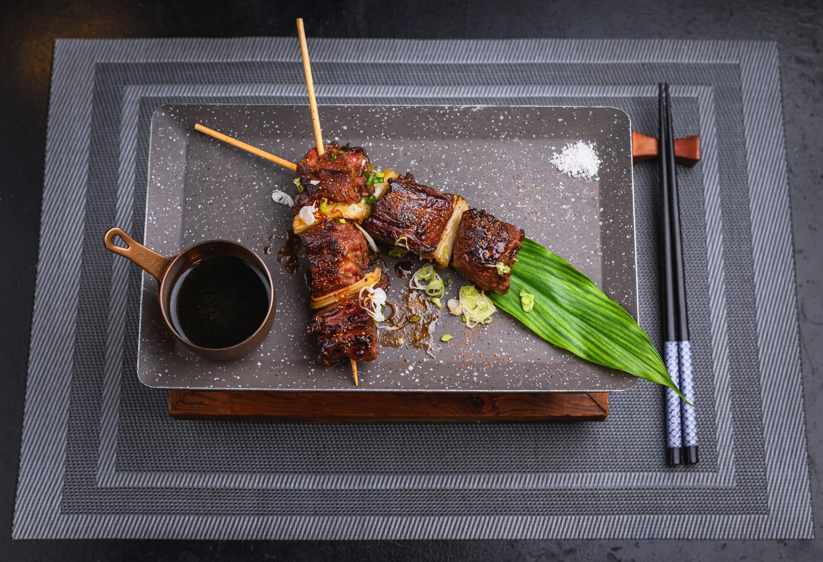 Broiled beef short ribs garnished with green onions, served with a side of dipping sauce, on a rectangular tray with a large green leaf, chopsticks, salt, and pepper.