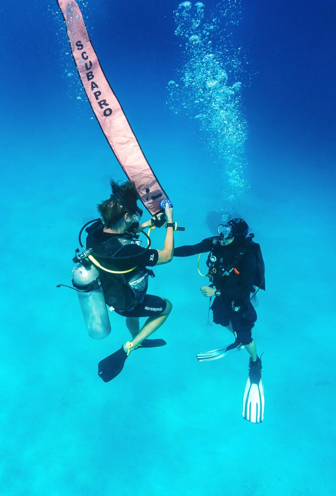 Two scuba divers underwater, with one handing a pink SUBARU banner to the other, surrounded by blue ocean water.