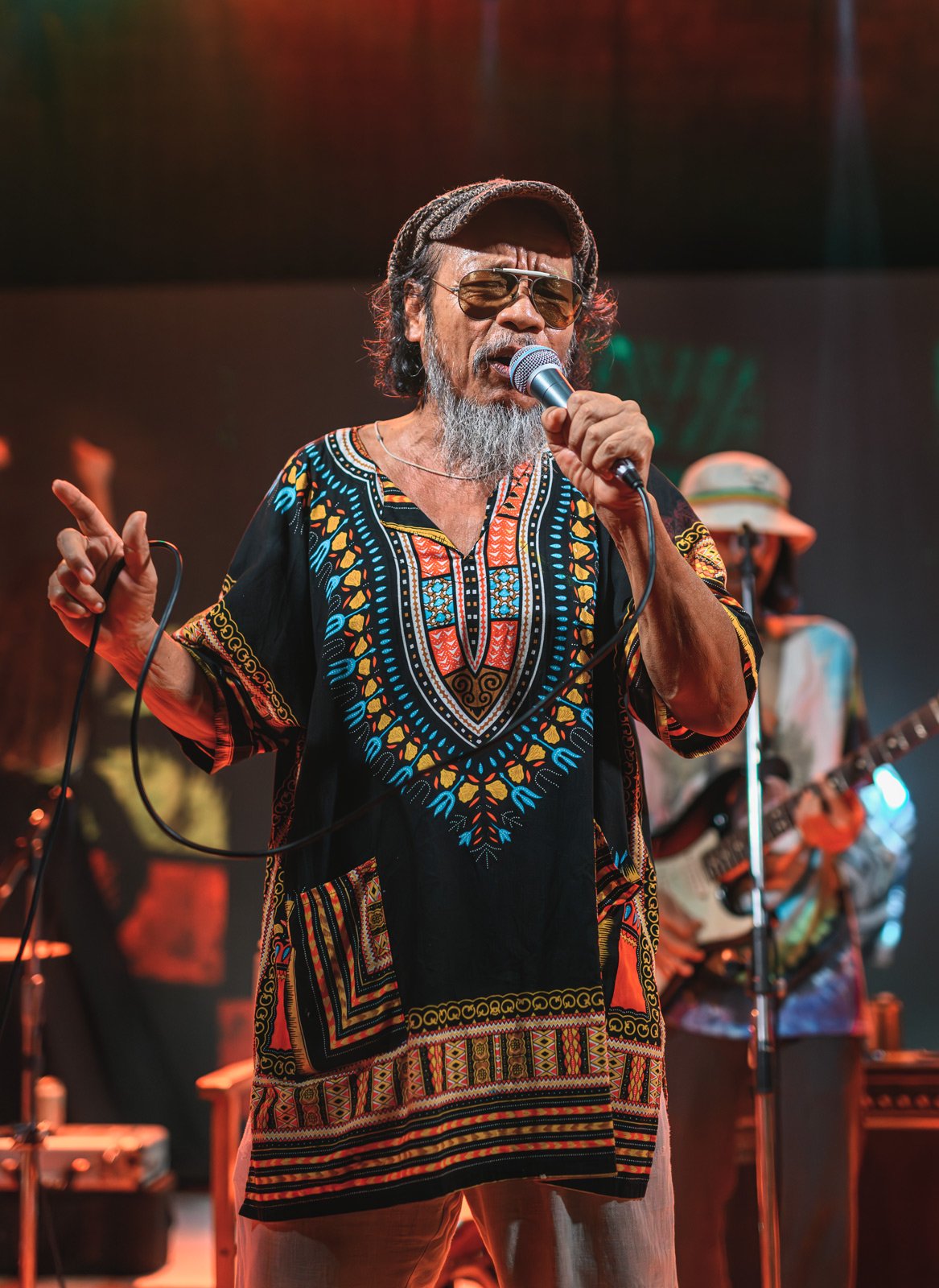 A man with a gray beard and glasses, wearing a colorful traditional shirt, holding a microphone and singing on stage. In the background, a woman in a hat is playing guitar.