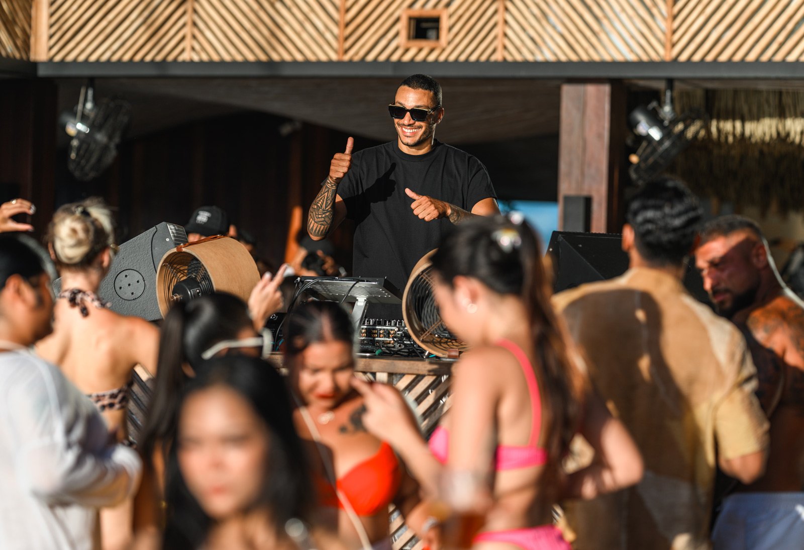 A DJ with tattoos and sunglasses smiling and giving a thumbs up while standing behind DJ equipment at a lively outdoor party with people dancing and enjoying the music.