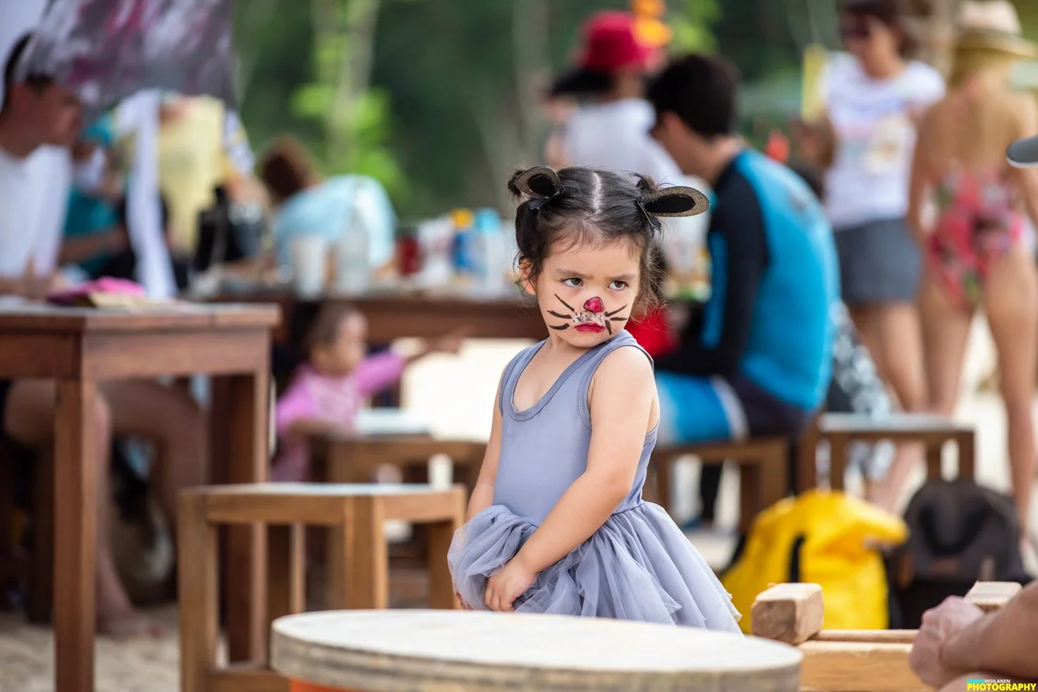 A young girl with face paint resembling a cat, including a pink nose and whiskers, stands with a serious expression at an outdoor gathering. She wears a gray dress and has her hair styled in two buns with cat ear accessories. In the background, sever