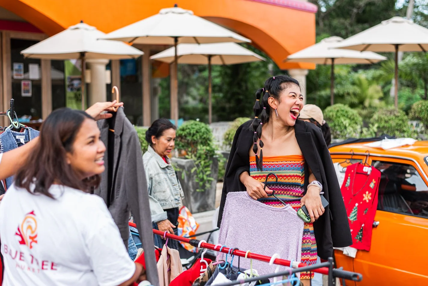 A group of women shopping outdoors at a flea market, with one woman in colorful clothing laughing and holding a small bag, while others browse clothes on a rack.