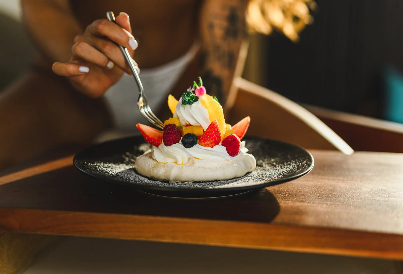 A person is eating a decorated fruit and whipped cream dessert on a black plate.
