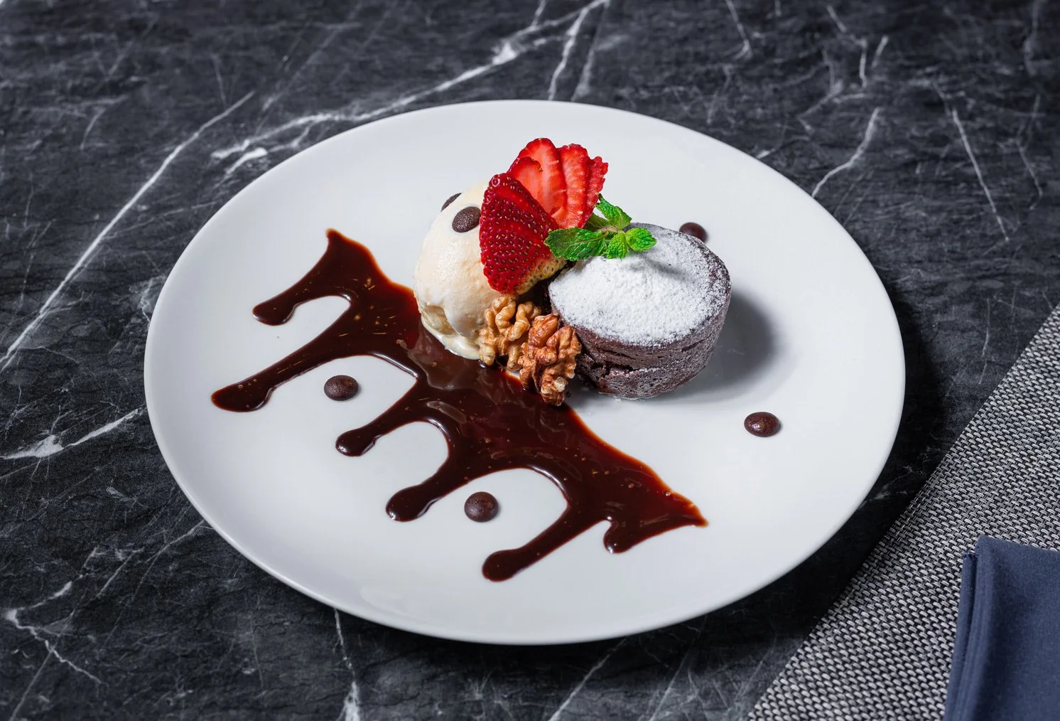 Chocolate lava cake with powdered sugar, garnished with sliced strawberries and mint leaves, served with a scoop of vanilla ice cream with chocolate drops, on a white plate with chocolate sauce decorations.