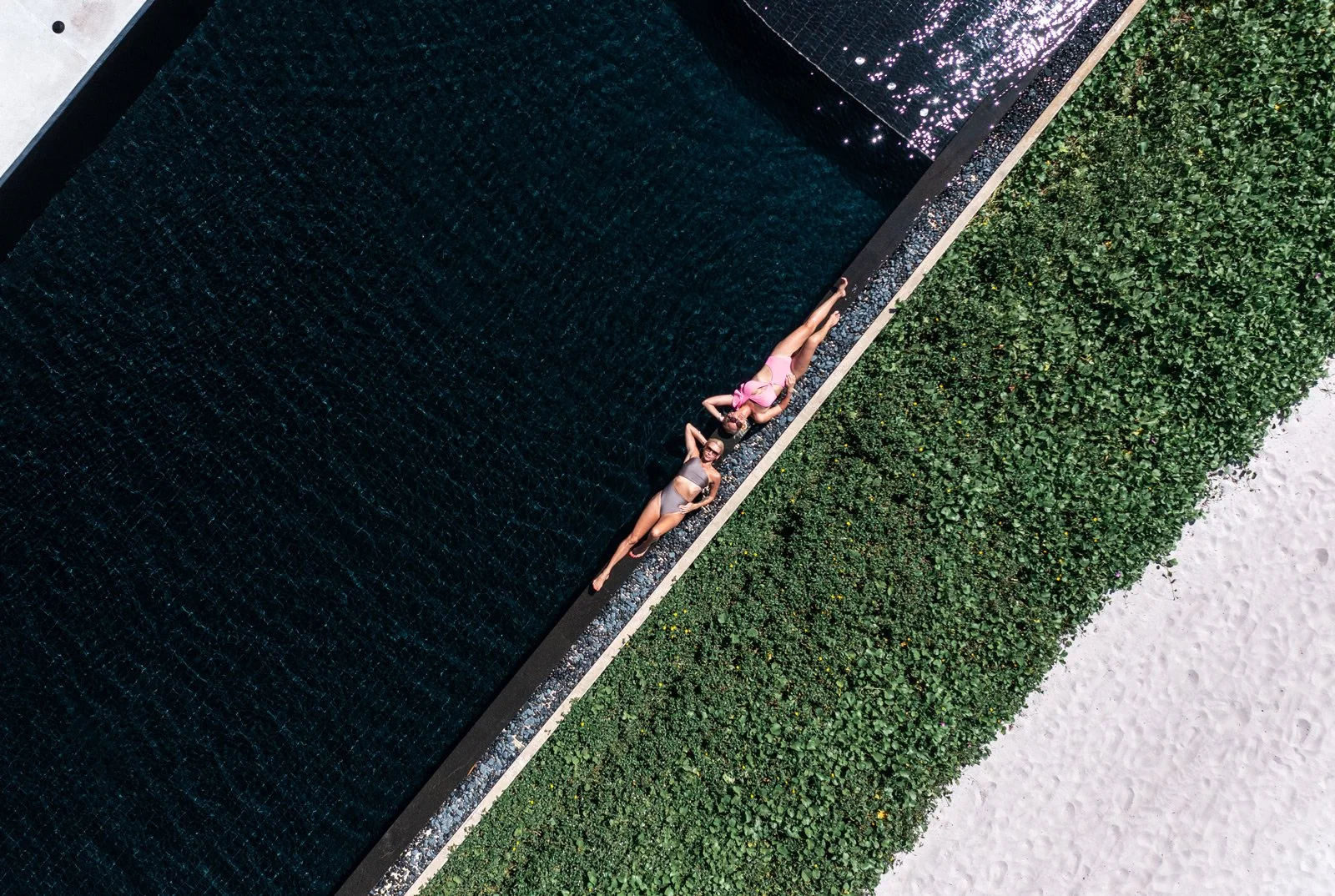 Two women in swimsuits lounging by a dark swimming pool, with one woman lying on her side and the other on her back, along a strip of green bushes and white sand.